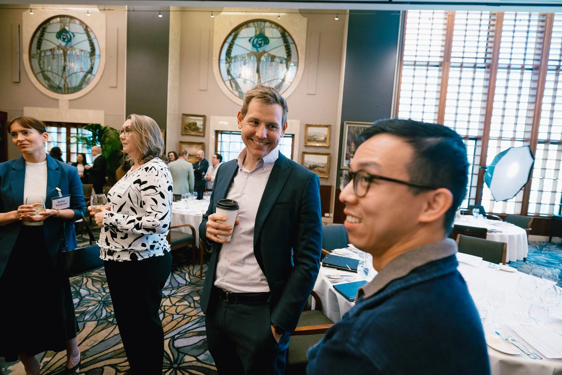 People socializing at a restaurant or banquet hall, engaged in conversation, holding drinks, with formal attire and a decorated interior.