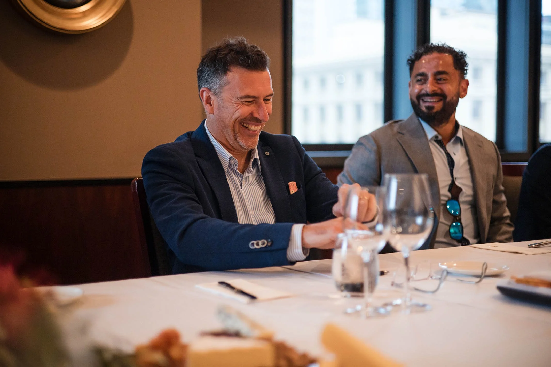 Two men in suits sitting at a banquet table, smiling and laughing, with wine glasses and dinnerware in front of them, in a well-lit restaurant or event space.