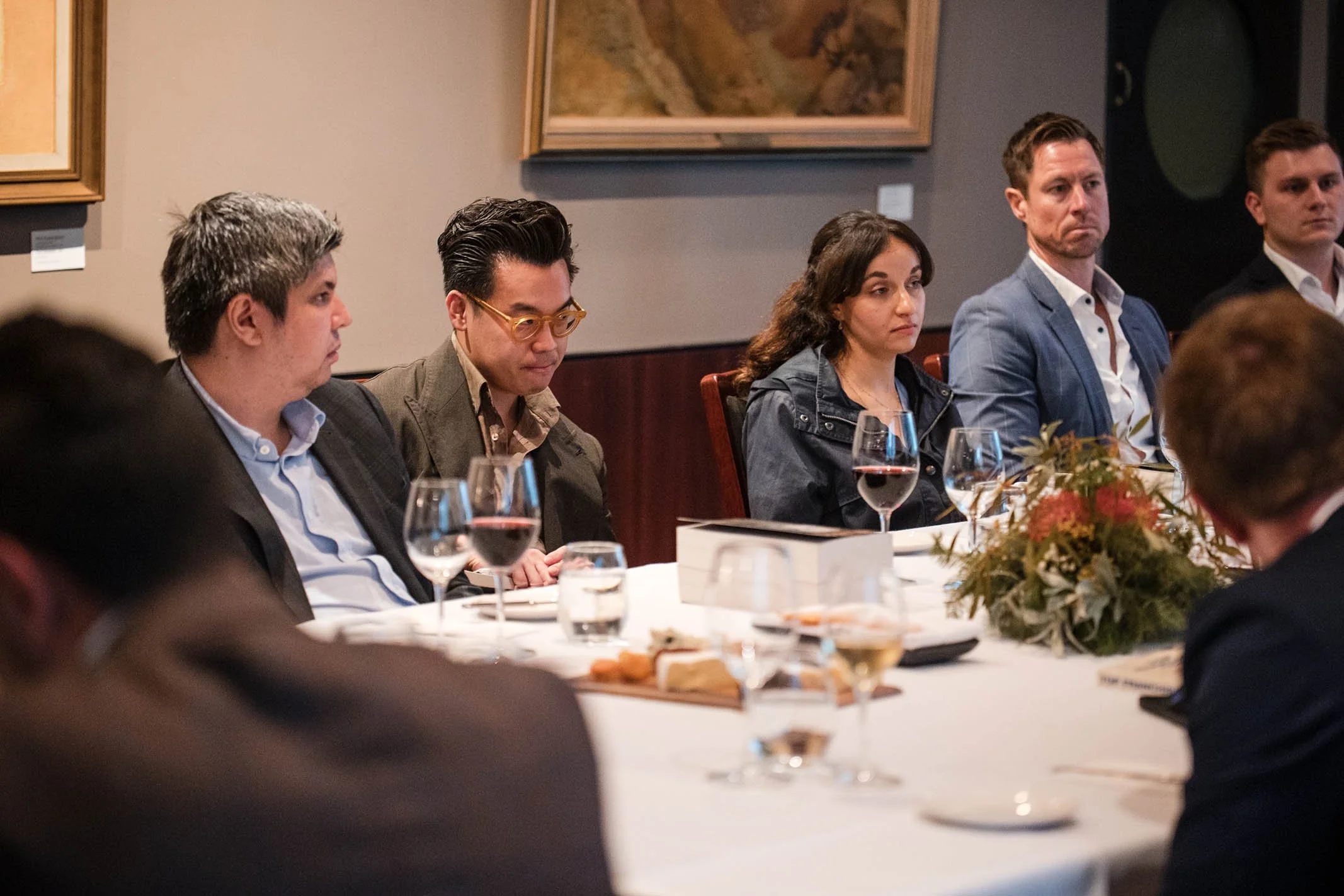 People sitting at a dinner table during a formal event, with glasses of wine and a floral centerpiece.
