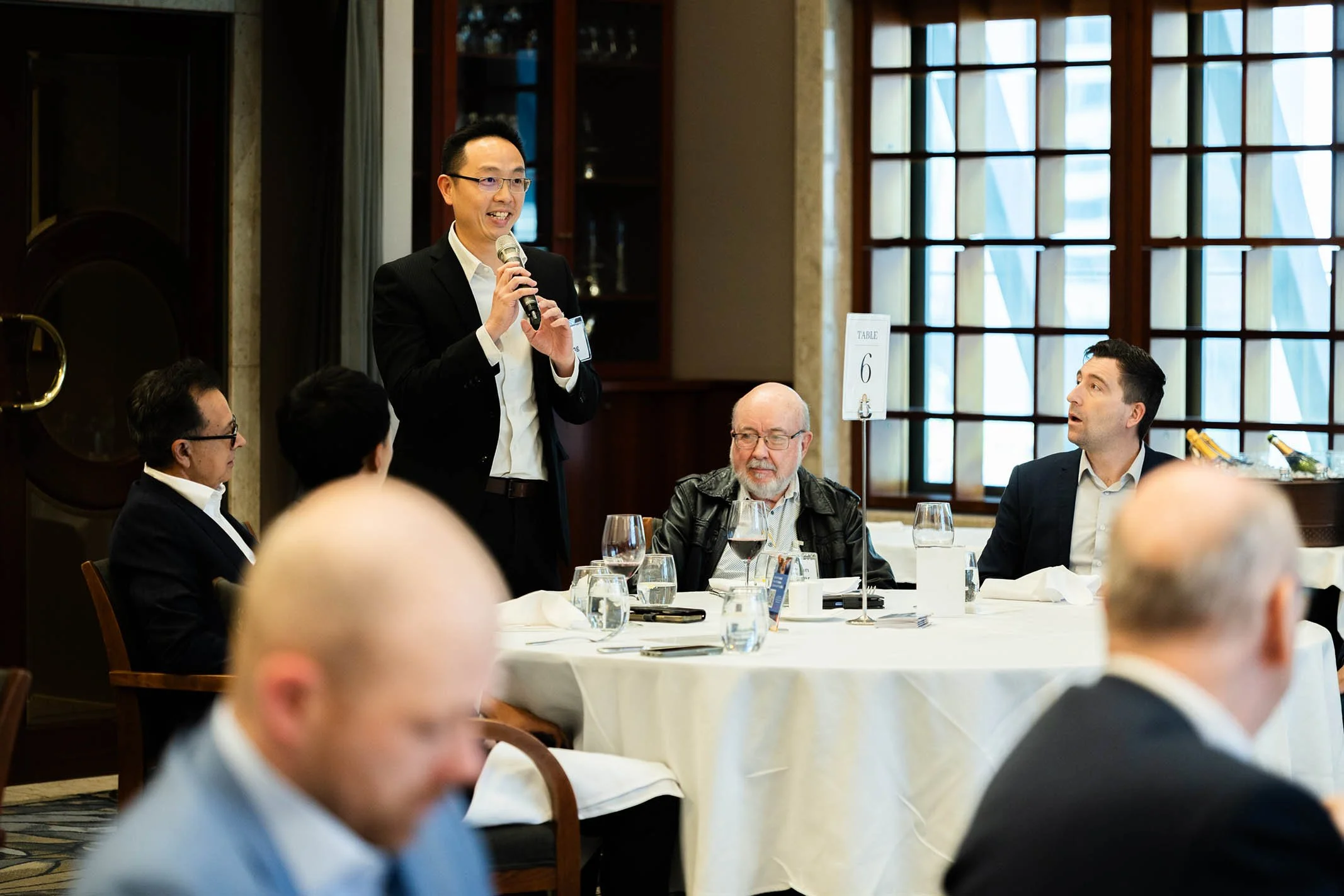A man in a black suit with glasses is speaking into a microphone at a round table during a formal event. Four other people sit around the table, listening. The table has water glasses, a menu, and a sign with the number 6. The background features lar
