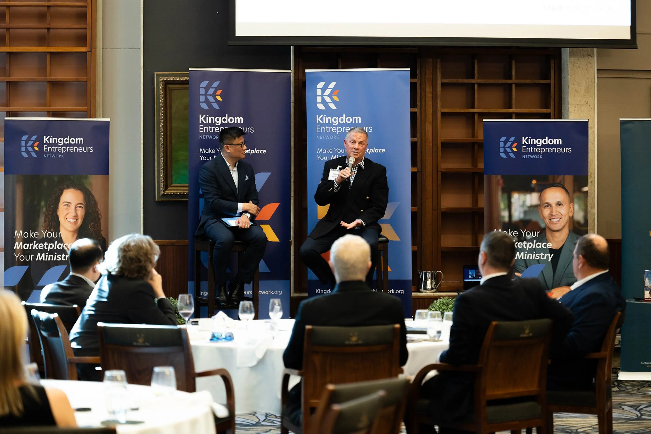 A professional conference or seminar with two men speaking on stage; one man is holding a microphone, and the other is seated, listening. The event is called 'Kingdom Entrepreneurs Network,' with banners displaying slogans and images of smiling indiv