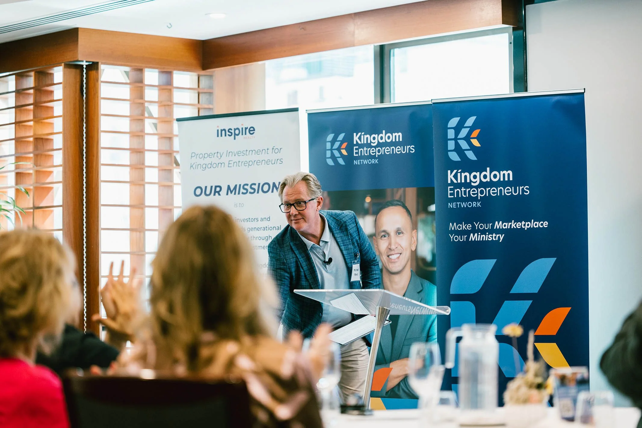 A man in a blue checked blazer and glasses speaking at a conference, with a lectern and an audience in the foreground. Behind him are banners from Kingdom Entrepreneurs Network and Inspire Property Investment for Kingdom Entrepreneurs.