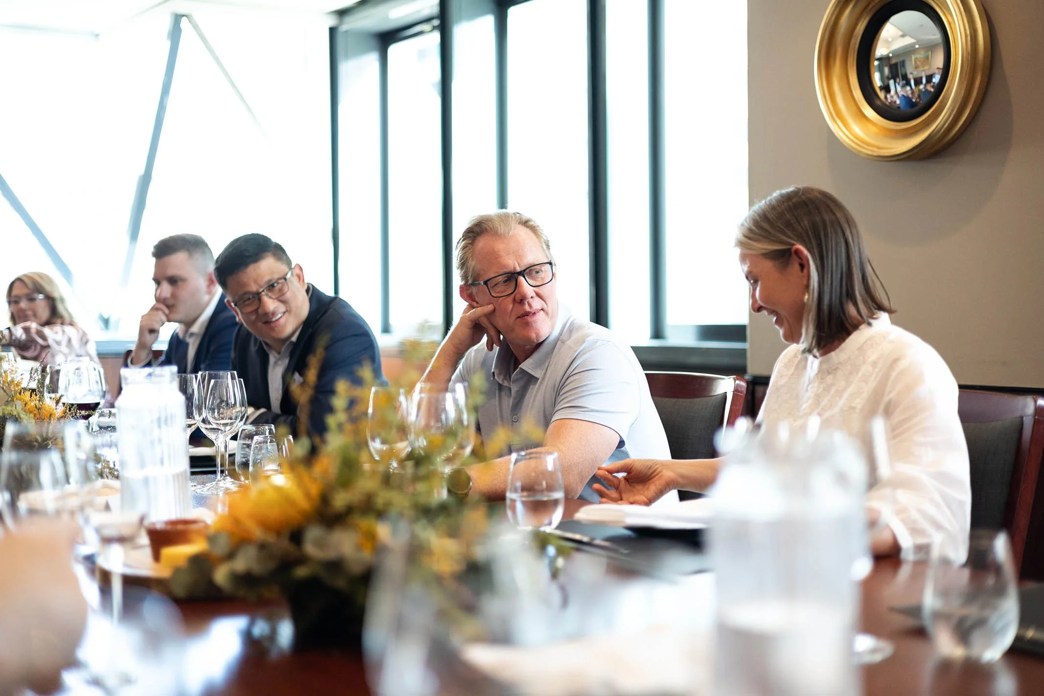 People sitting at a long dining table, engaged in conversation during a formal meal in a well-lit room with large windows and modern decor.