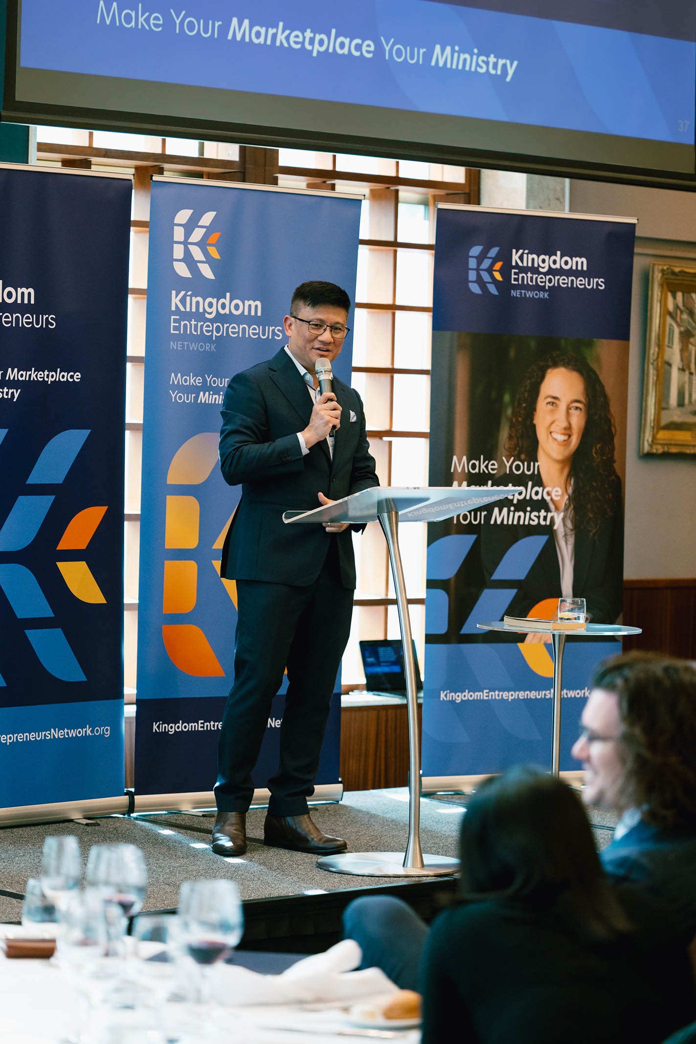 A man in a dark suit and glasses speaking into a microphone at a podium during a presentation or conference, with banners behind him displaying the logo and name "Kingdom Entrepreneurs Network" and the slogan "Make Your Marketplace Your Ministry".