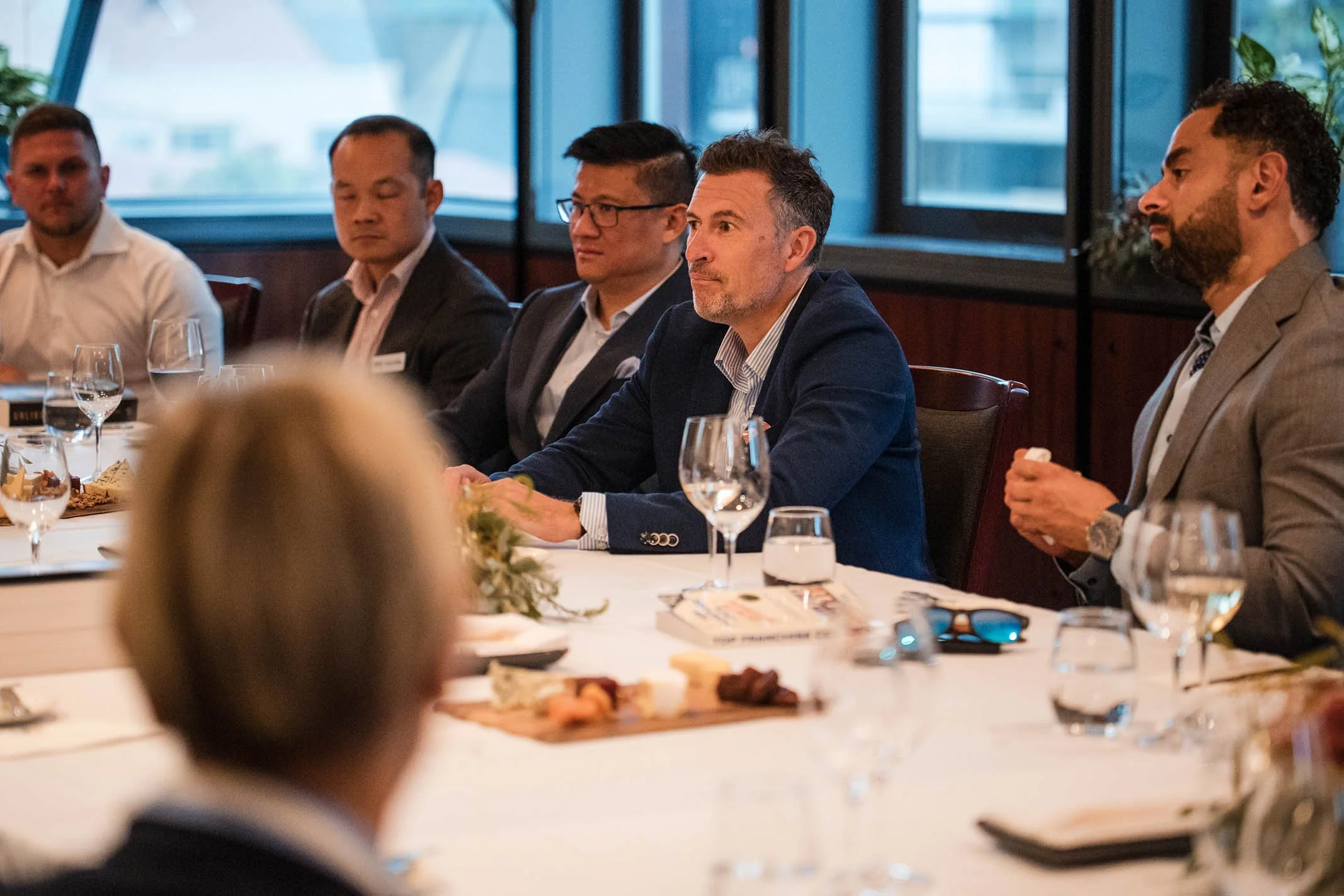 A group of professionally dressed people attending a formal meeting or conference around a table with glasses, notebooks, and plates of snacks in a well-lit room with large windows.