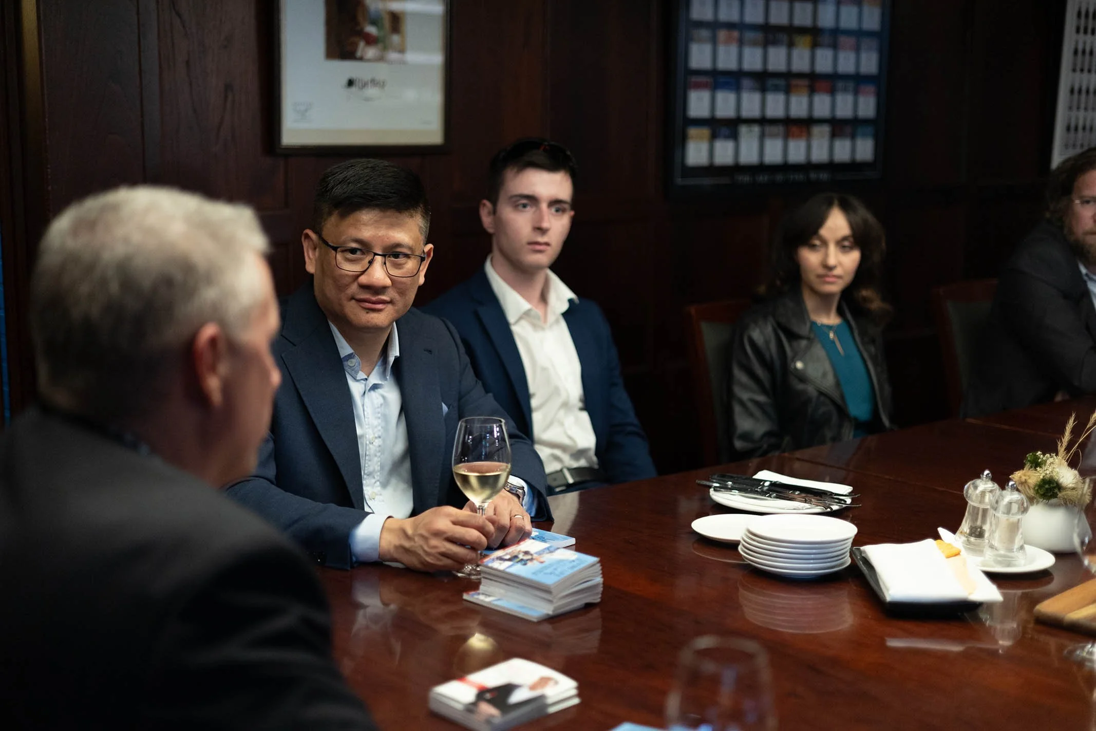 People sitting at a conference table during a formal meeting, with plates, glasses, and napkins arranged on the table.