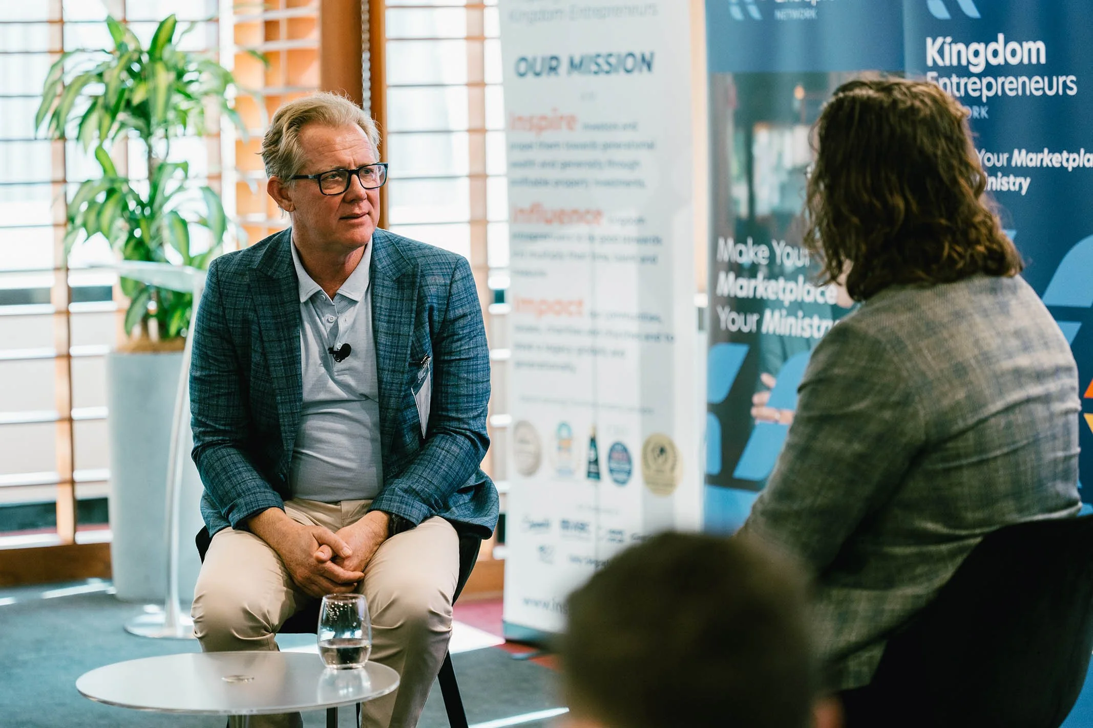 Two people having a conversation in a professional setting. The man on the left has gray hair, glasses, and is wearing a checkered blazer with a white shirt, while the woman on the right, with curly hair, is dressed in a gray blazer. Behind them are 