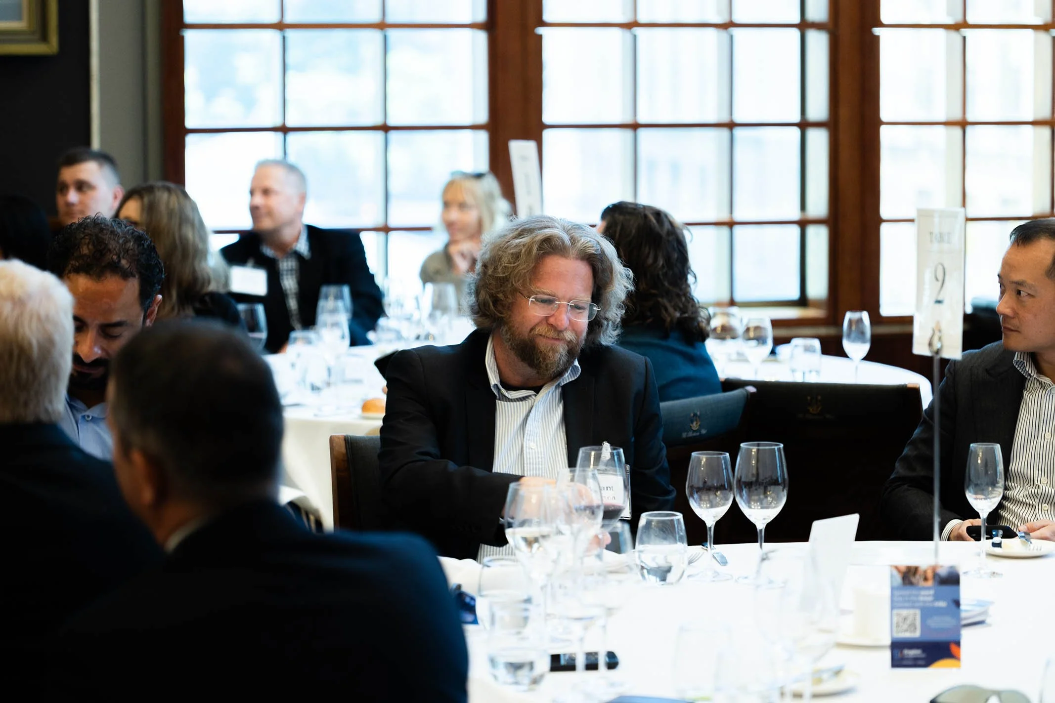 People sitting at round tables in a well-lit room with large windows, engaging in conversation during a formal event or conference, with glasses and table settings visible.