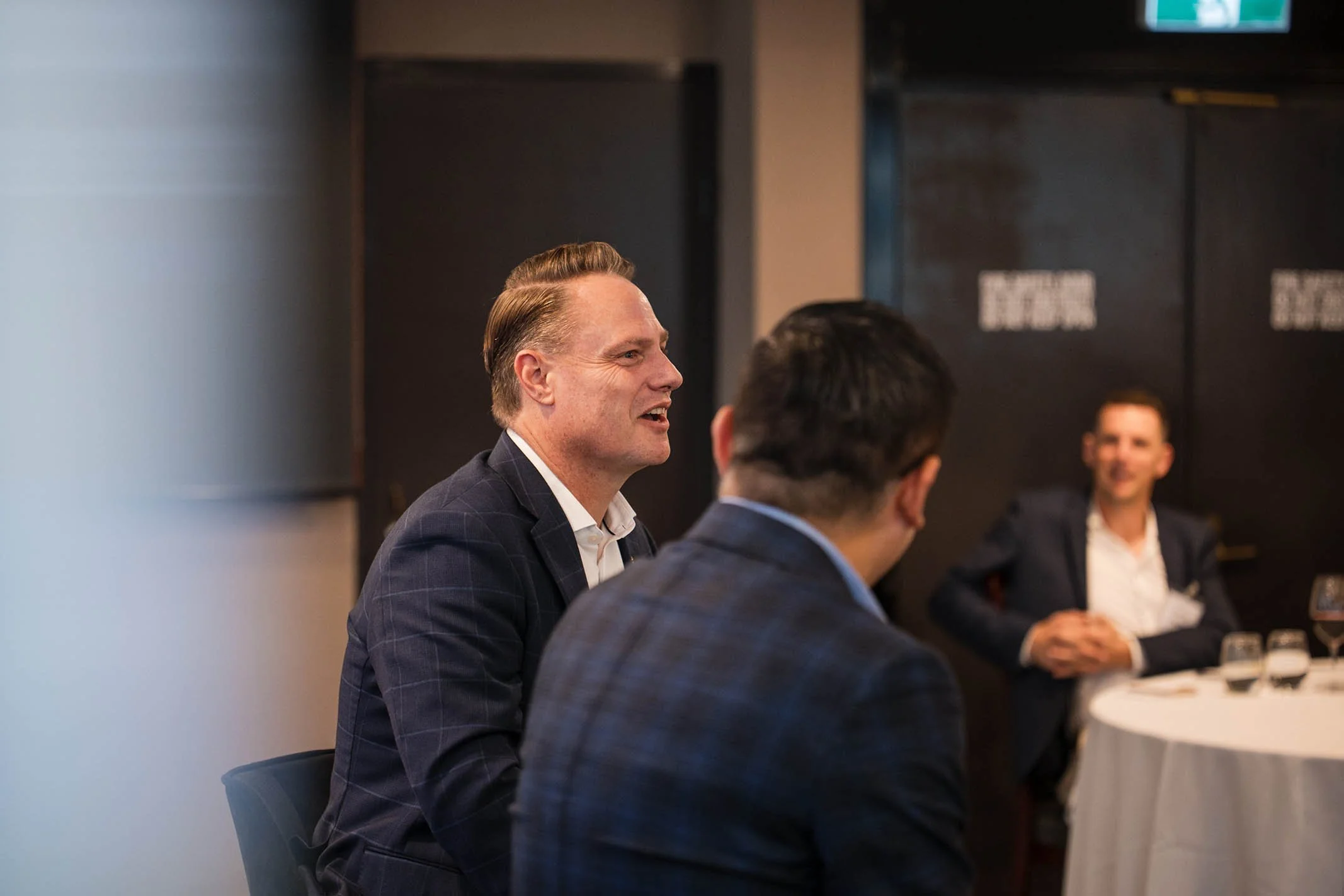 Three men in suits sitting at a round table engaging in conversation at a formal event.