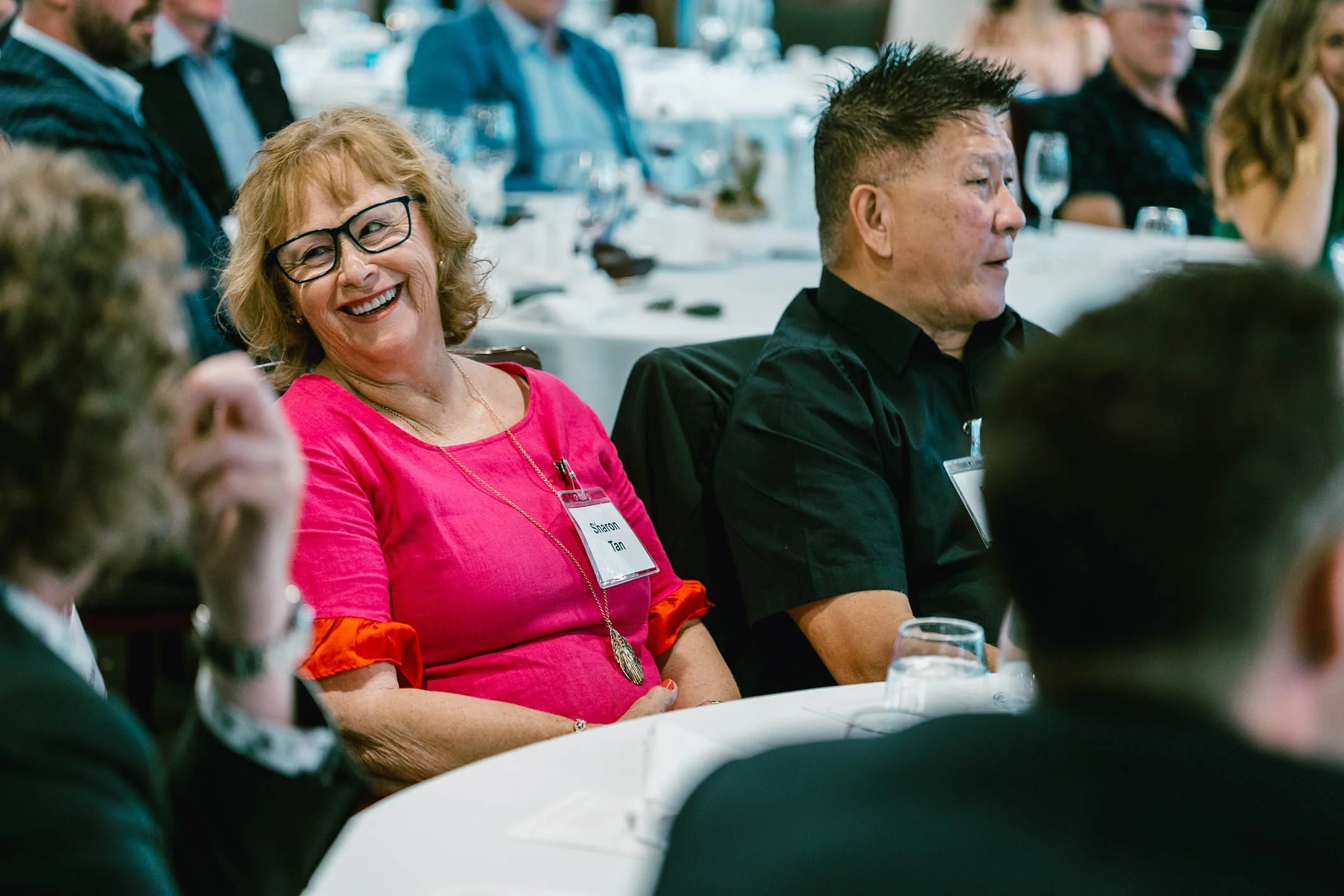A woman with curly blonde hair, glasses, and a bright pink dress is smiling and laughing at a social or professional event. She has a name tag that reads 'Sharon Tan' and is sitting at a table with other people in a banquet or conference setting.