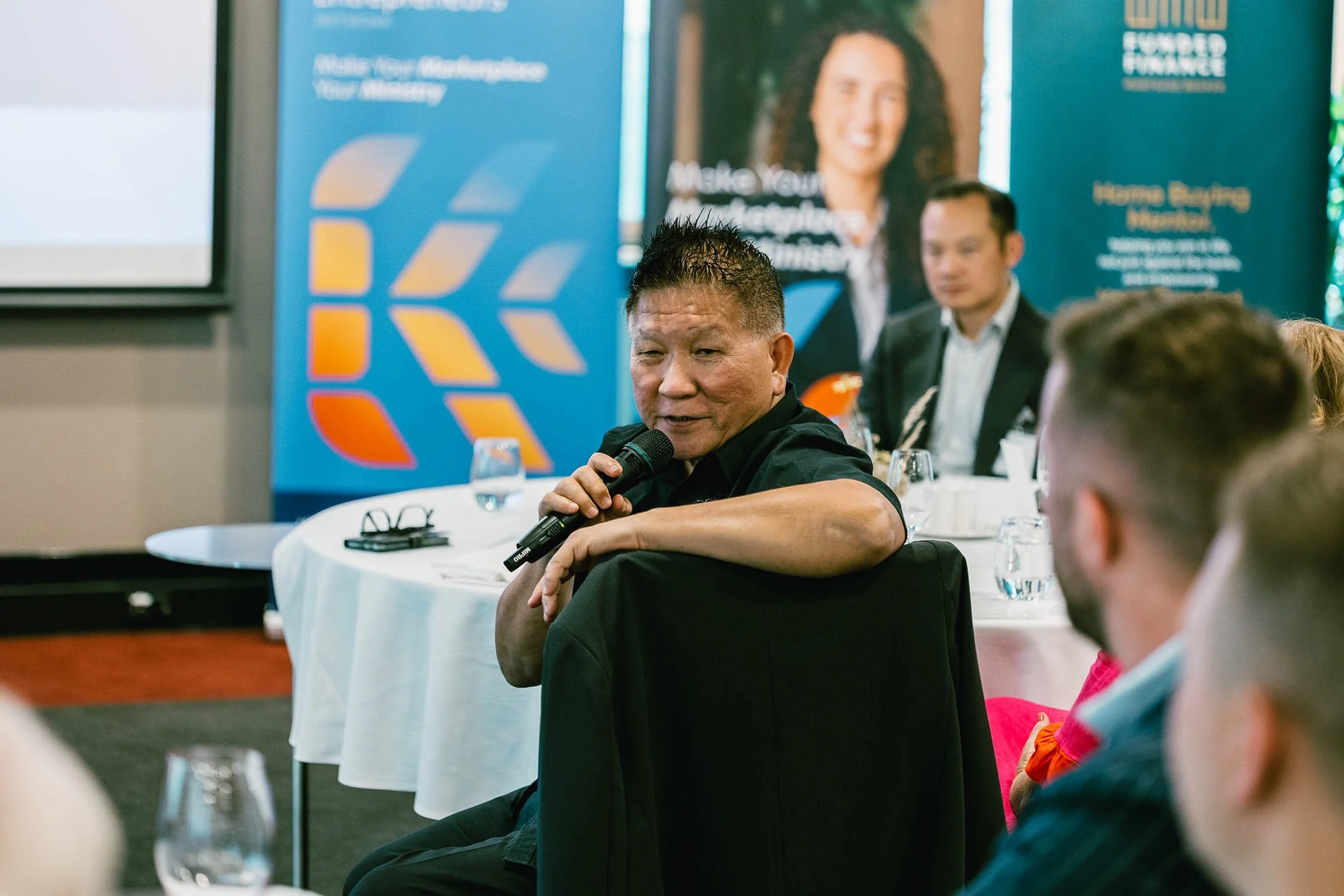 A man with short dark hair in a black shirt is holding a microphone and speaking at a conference, seated at a round table with water glasses. Behind him, there are other attendees, a large blue banner with text, and a projected screen.