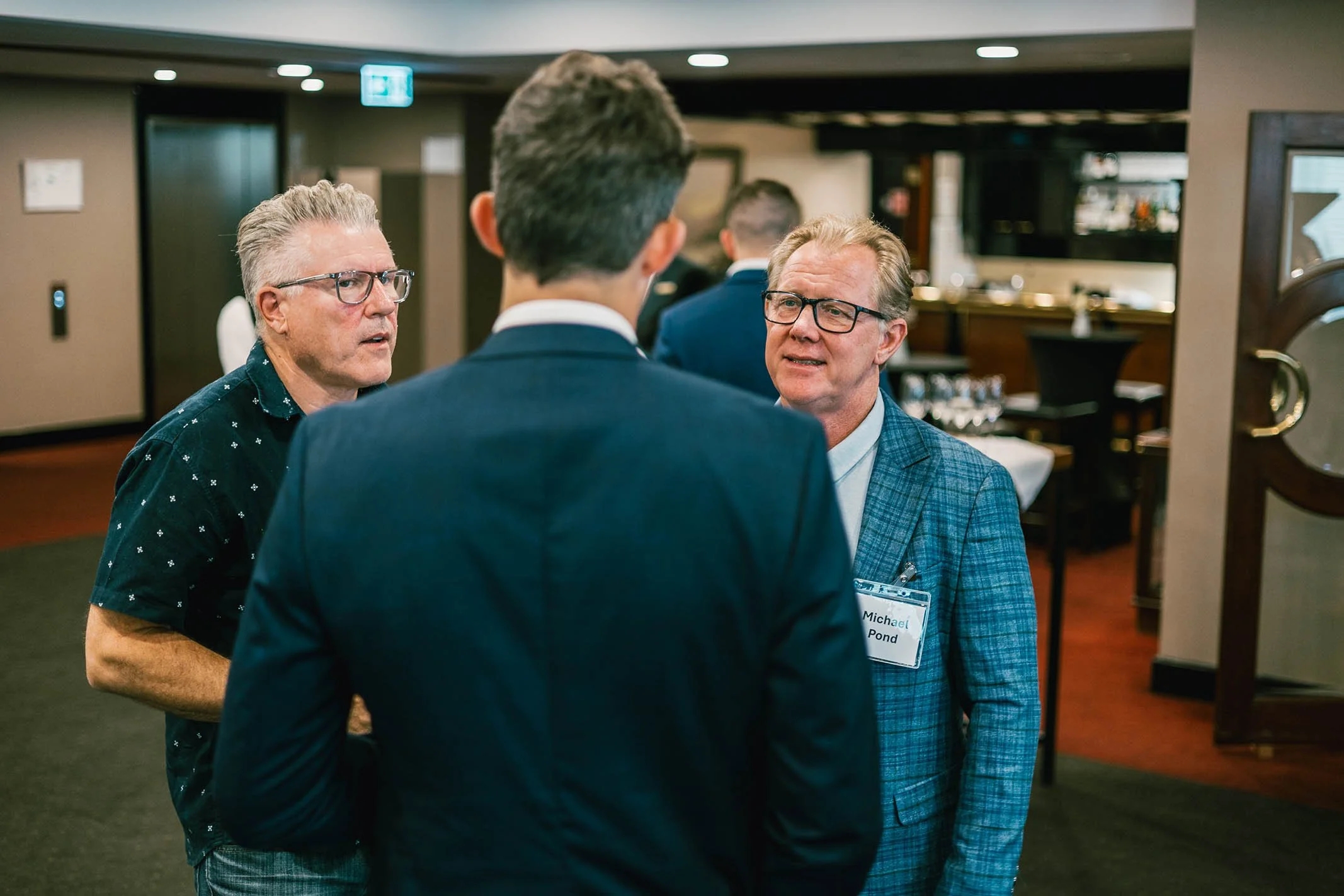 Three men in conversation at a formal event in an indoor venue with a bar and dining area in the background.