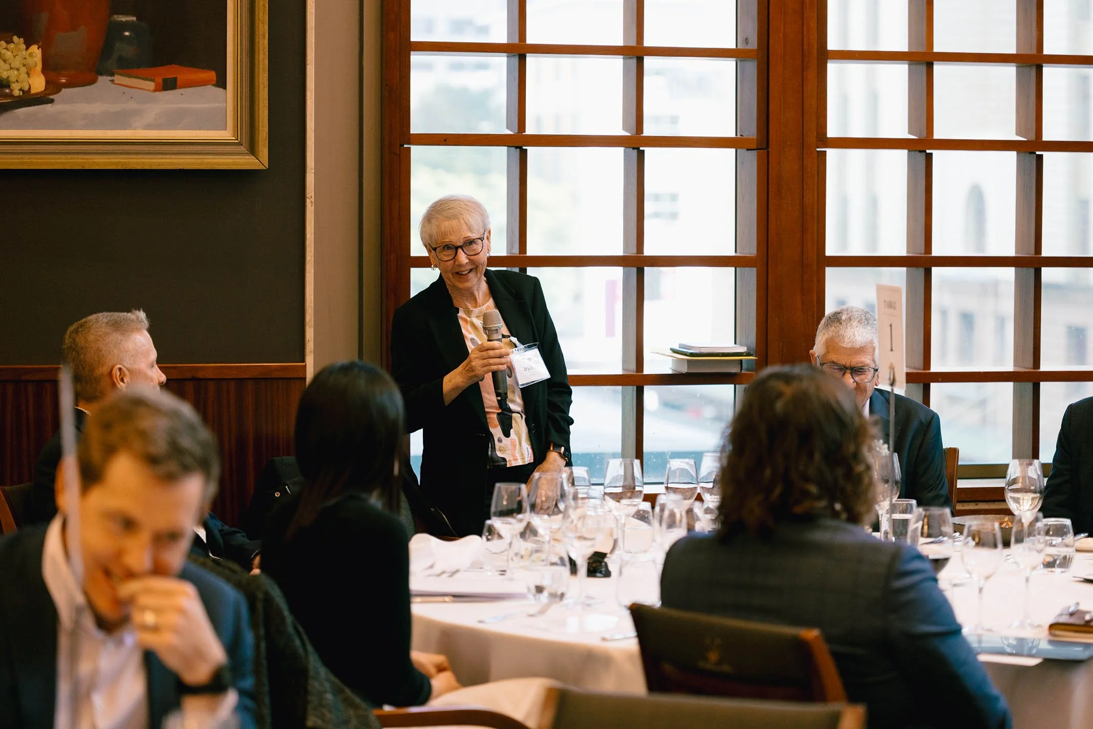 An elderly woman standing and speaking into a microphone at a banquet table during a formal gathering.