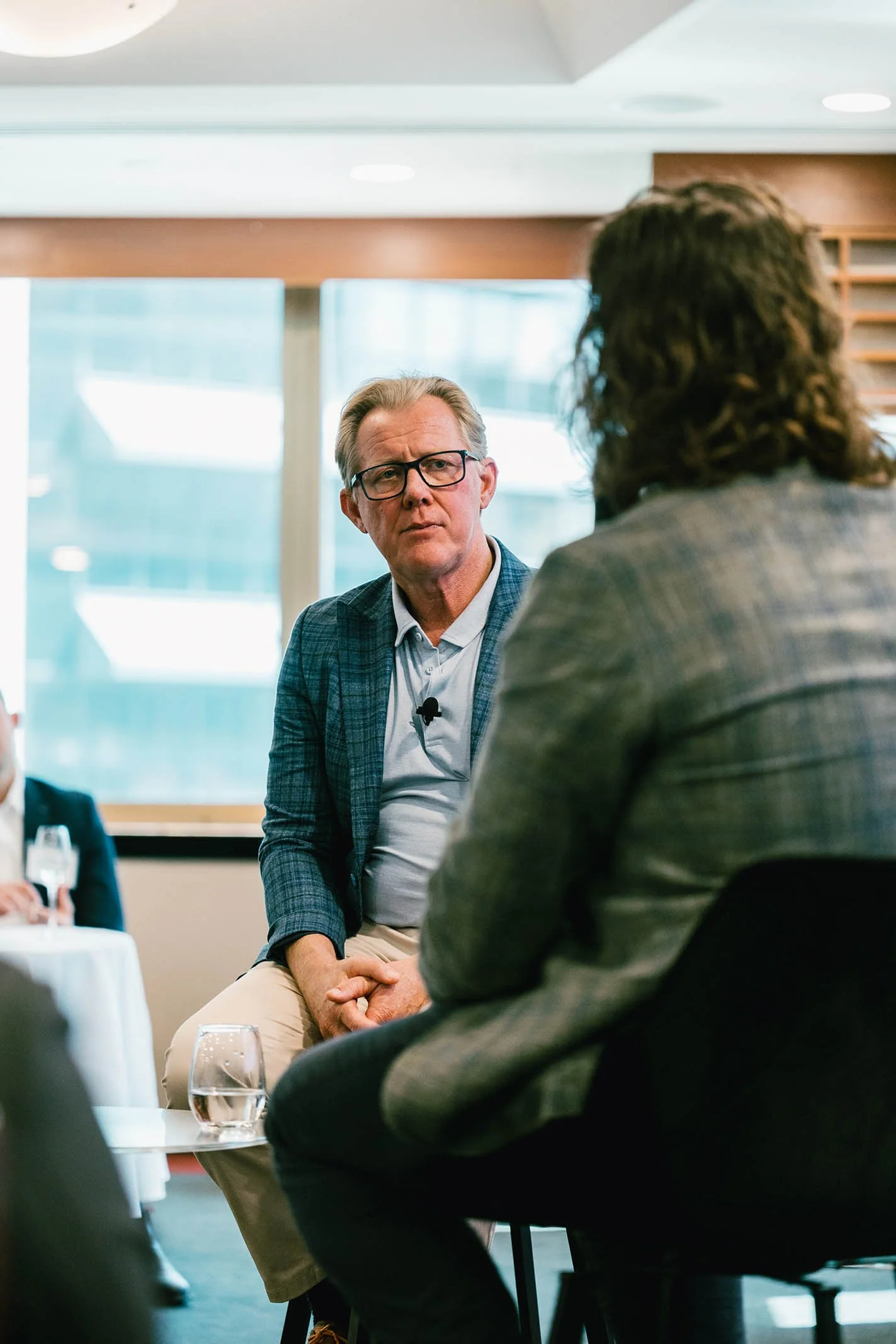Two men are engaged in a conversation during a professional event or panel discussion in a bright, modern room with large windows.