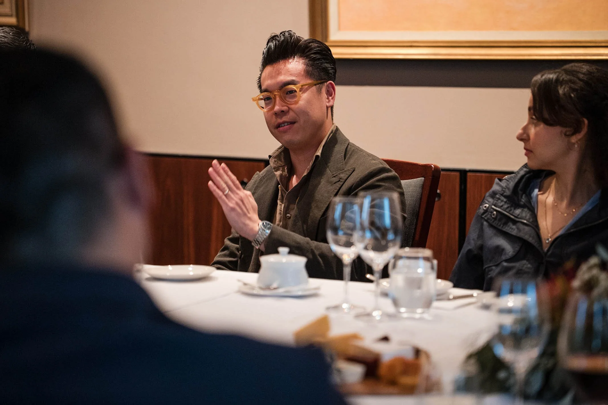 A man with glasses and a woman seated at a dining table engaged in conversation during a meal.