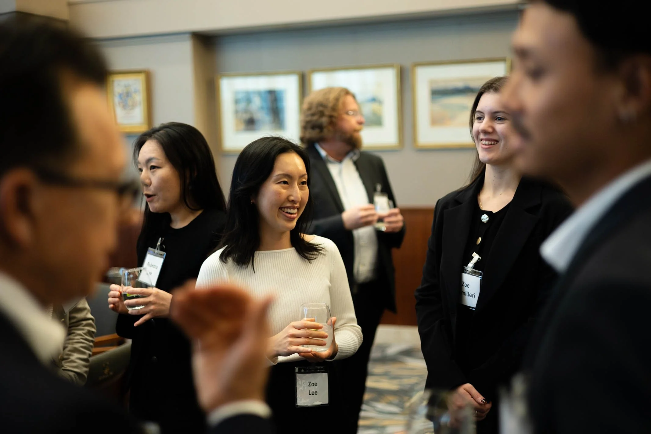 Group of diverse young professionals talking and smiling at a networking event, holding drinks, with artwork on the wall in the background.