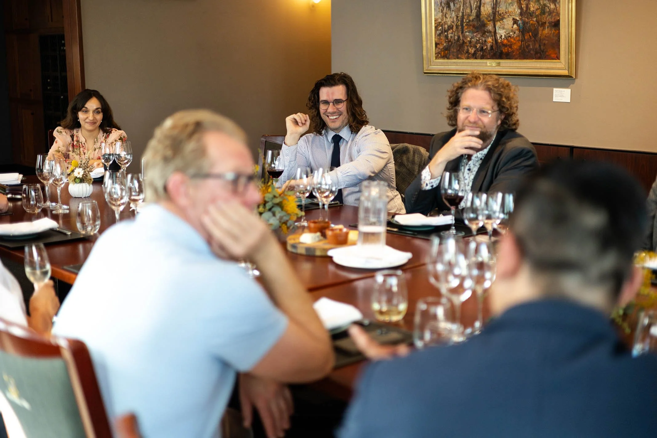 People sitting around a dining table, enjoying a gathering with wine glasses, flowers, and food, in a room with a painting on the wall.