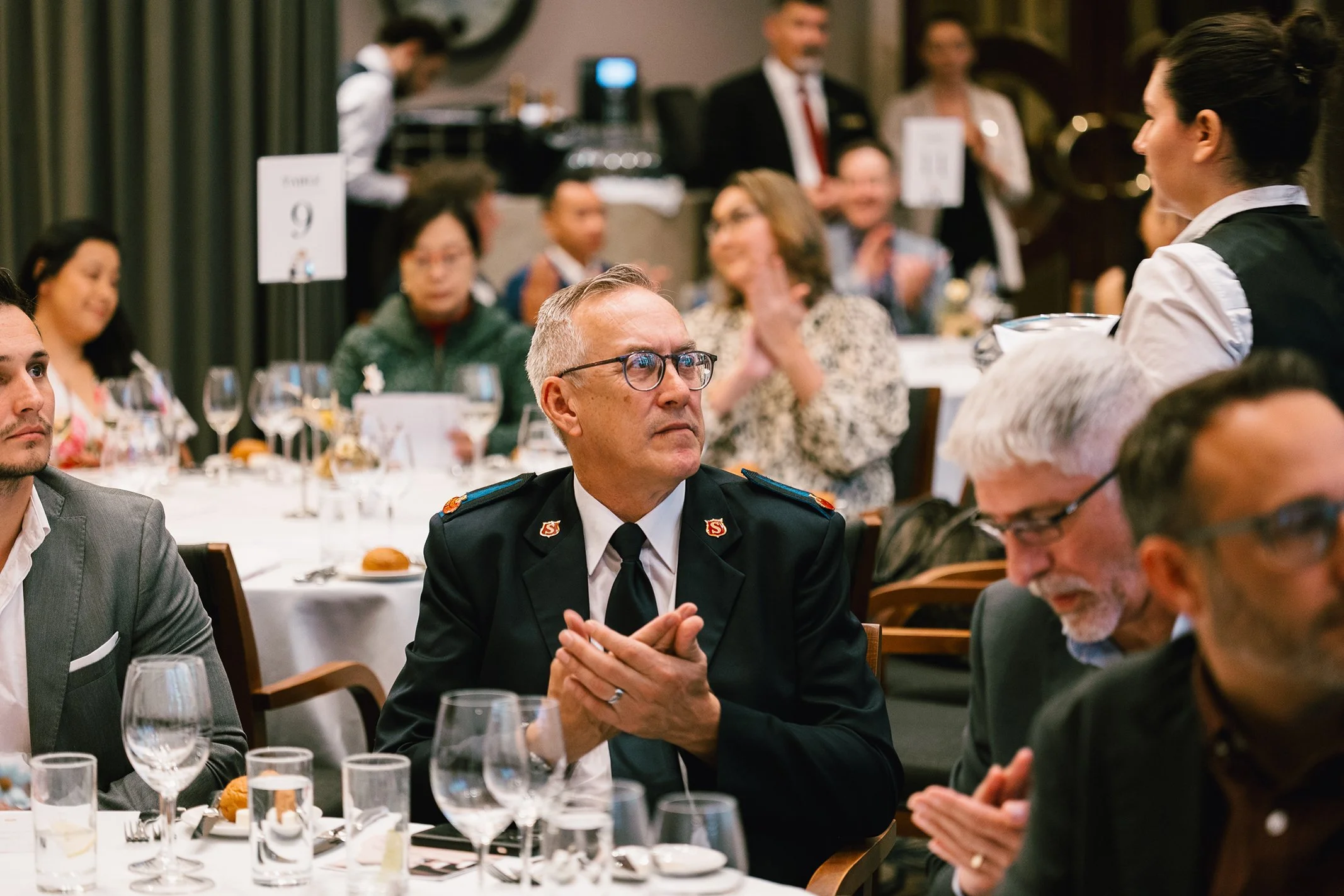 Man in military uniform with glasses sitting at a banquet table during an event, surrounded by other guests in formal attire.