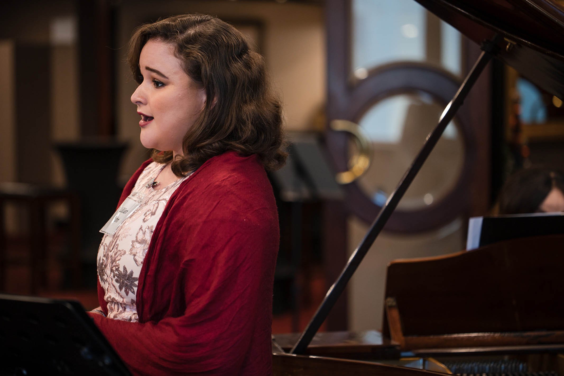A woman with brown wavy hair singing at a piano in a room with wooden decor and a large round mirror in the background.