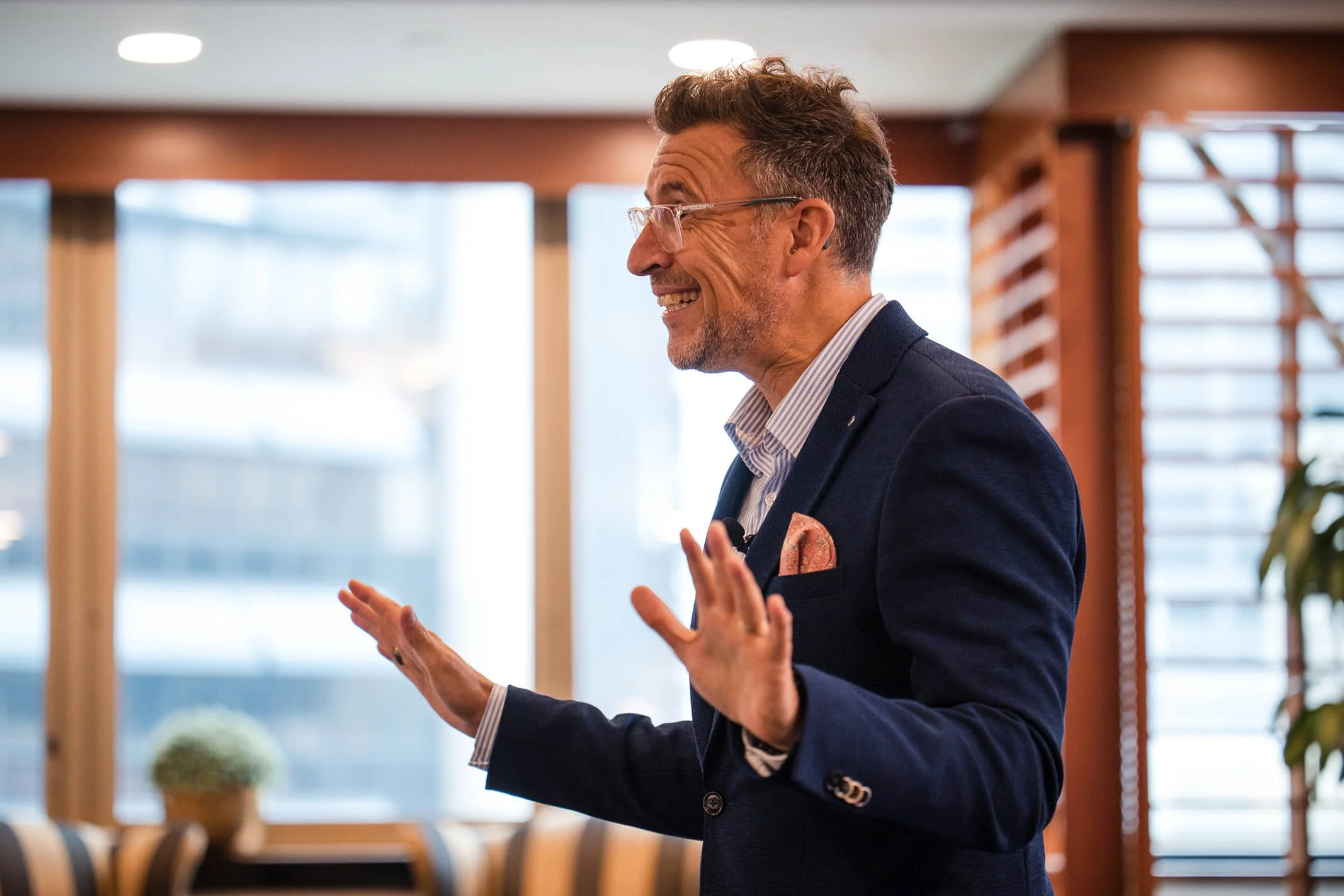 Smiling man with glasses, wearing a business suit, gesturing with his hands while speaking in a well-lit indoor setting.