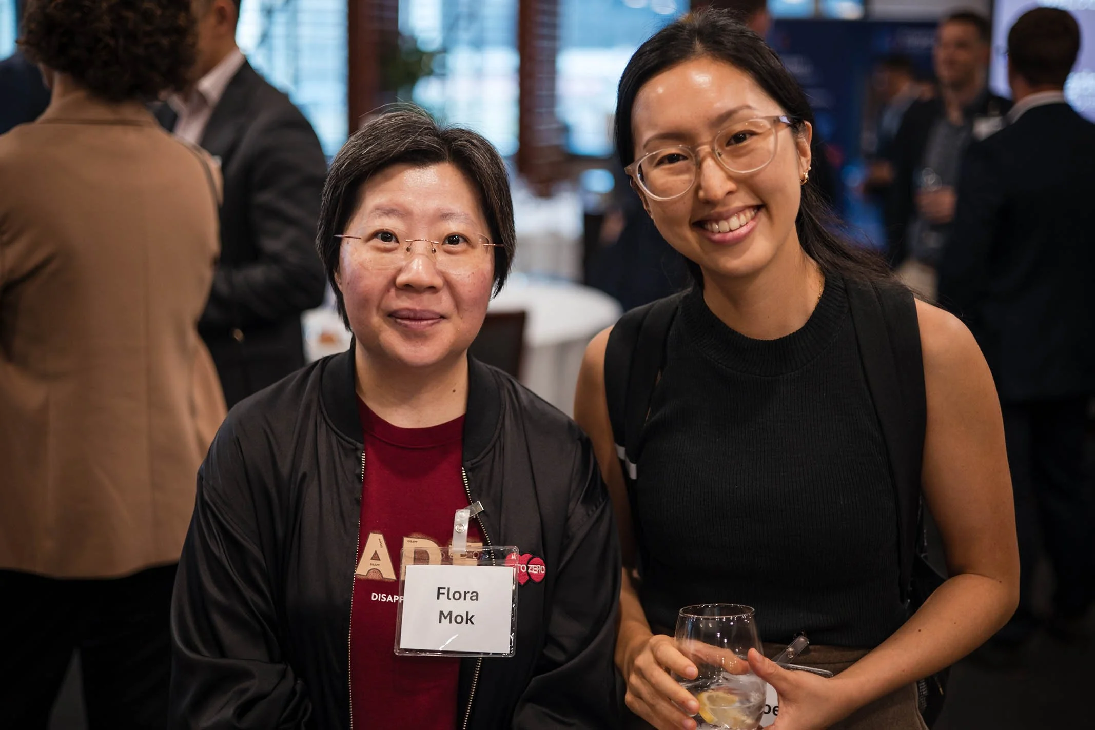 Two women standing together at a social event, smiling at the camera. One woman is wearing a black jacket and glasses, with a name tag that reads 'Flora Mok'. The other woman is wearing a black sleeveless top and glasses, holding a glass of water wit