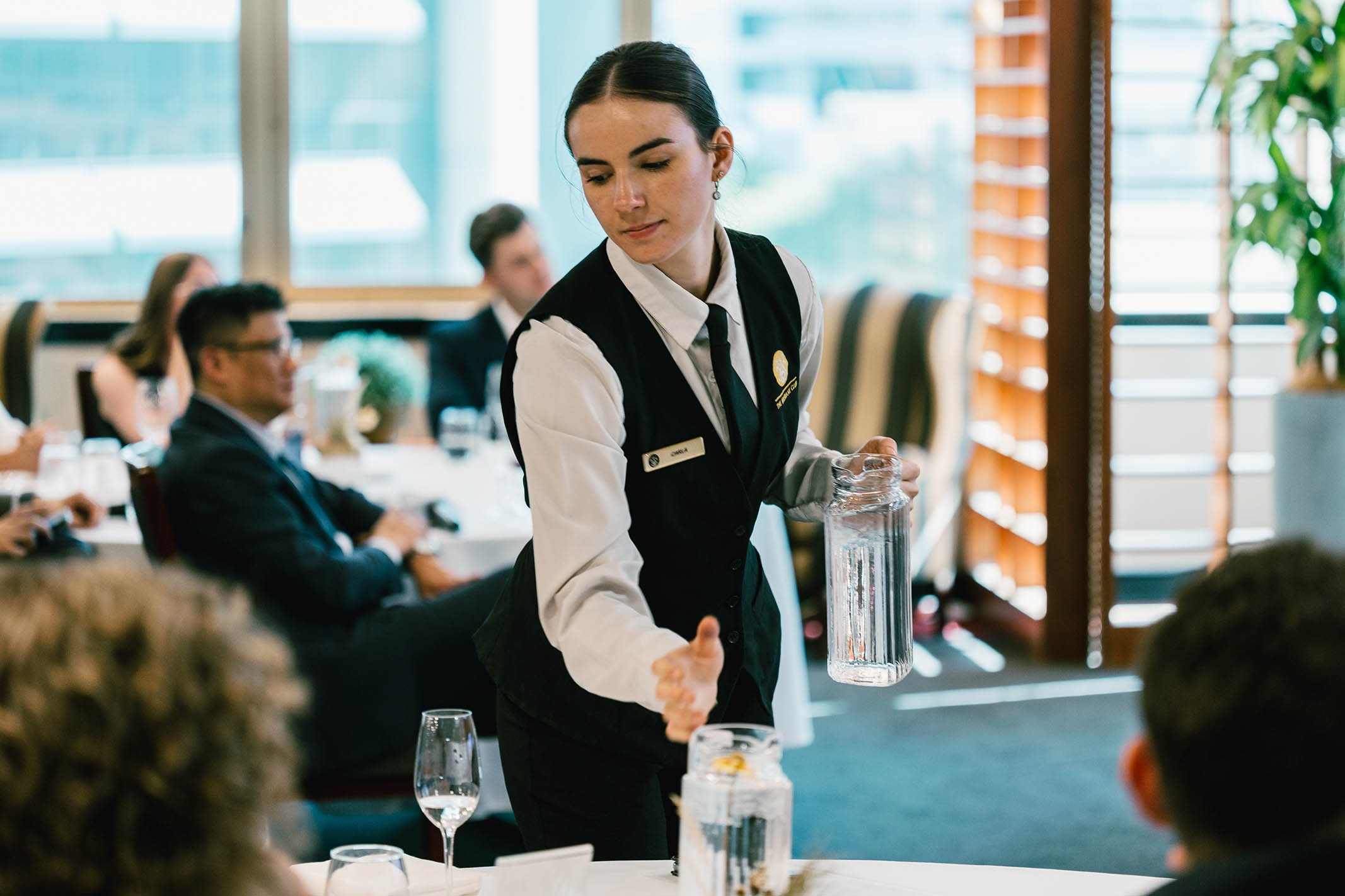 A young woman wearing a formal vest and white shirt serving water from a glass pitcher to guests at a dining event in a bright room with large windows and seated audience.