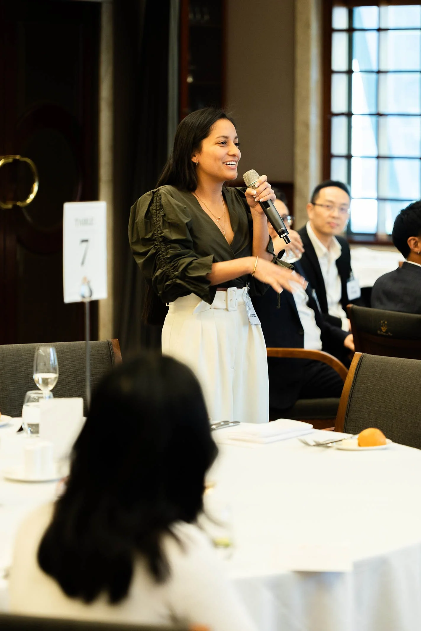 A woman standing and speaking into a microphone during a formal event or conference, with seated attendees in the background and a table with drink glasses and a plate of fruit in the foreground.