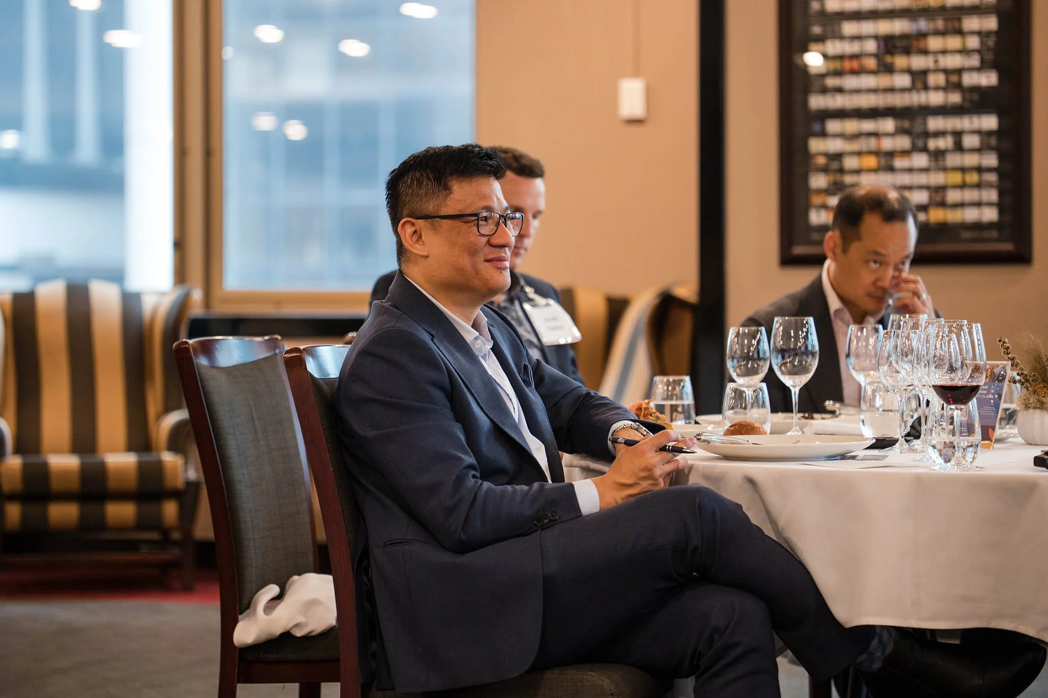 Business professionals at a formal dining event, seated at a table with wine glasses, plates, and silverware, in a well-lit indoor venue.