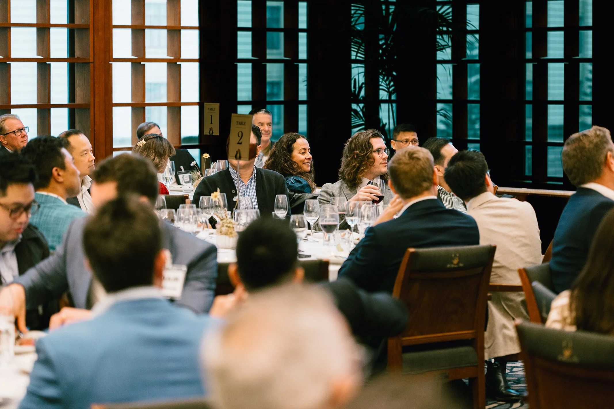Audience seated at round tables during a formal event or conference, with several drinks on the tables, and a large window with wooden grid design in the background.