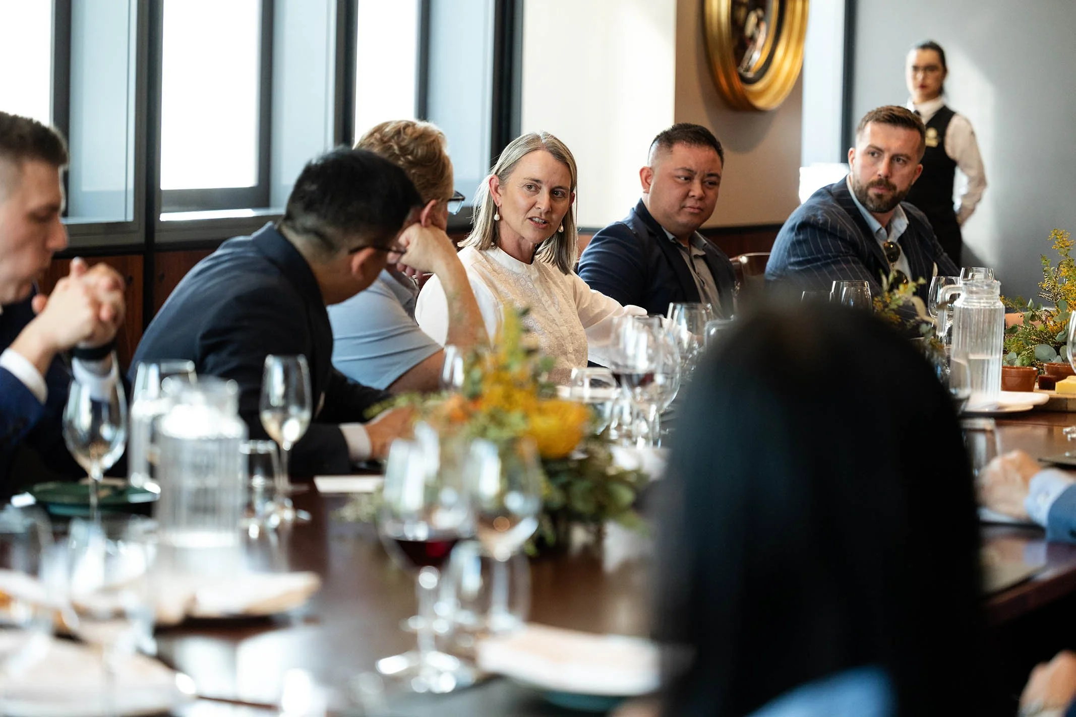 A group of people sitting at a long table during a meeting or conference, with a woman speaking and others listening, with flowers, glasses, and pitchers on the table, and a woman standing in the background.