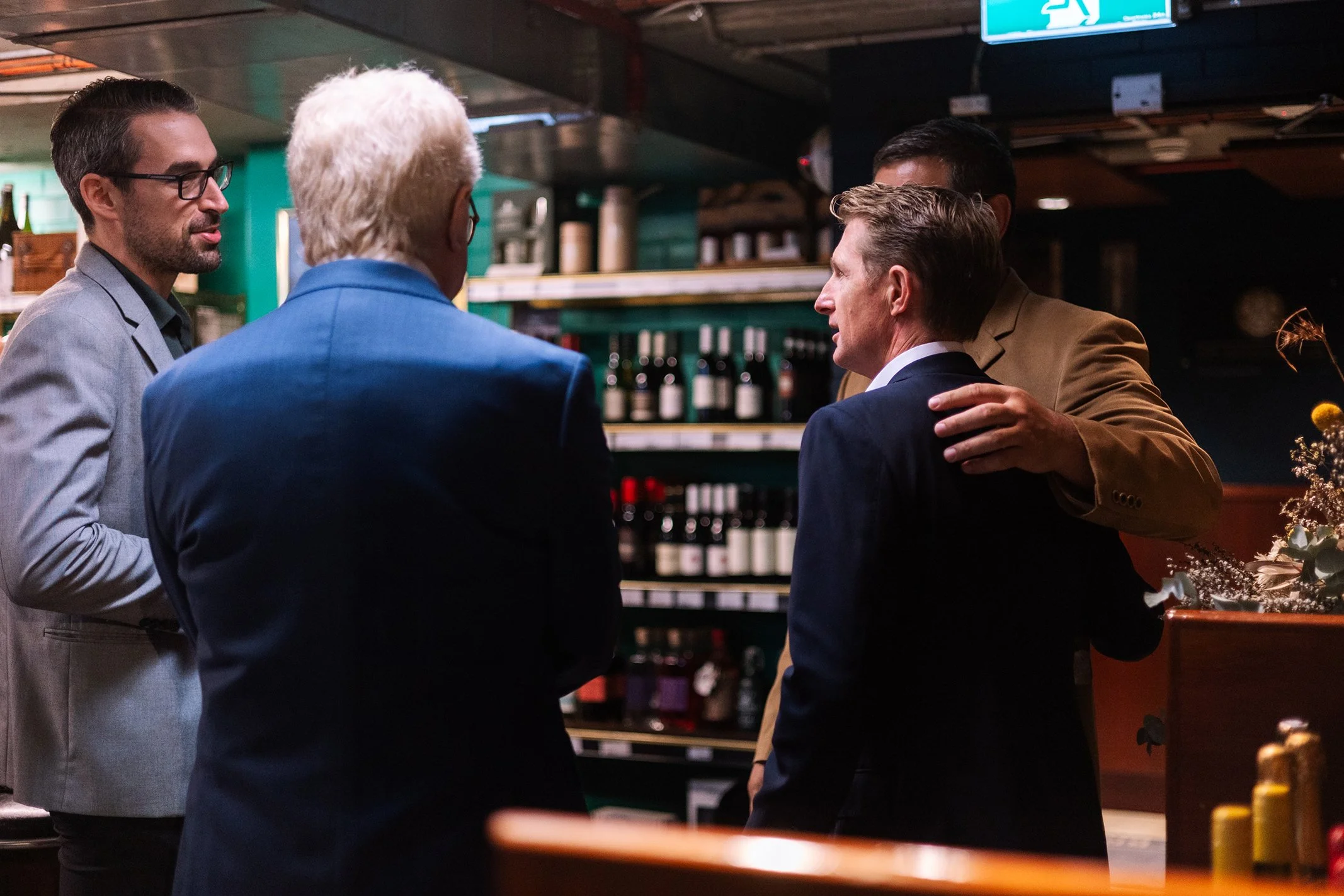 Four men in business attire engaged in a serious conversation in a wine shop or bar.