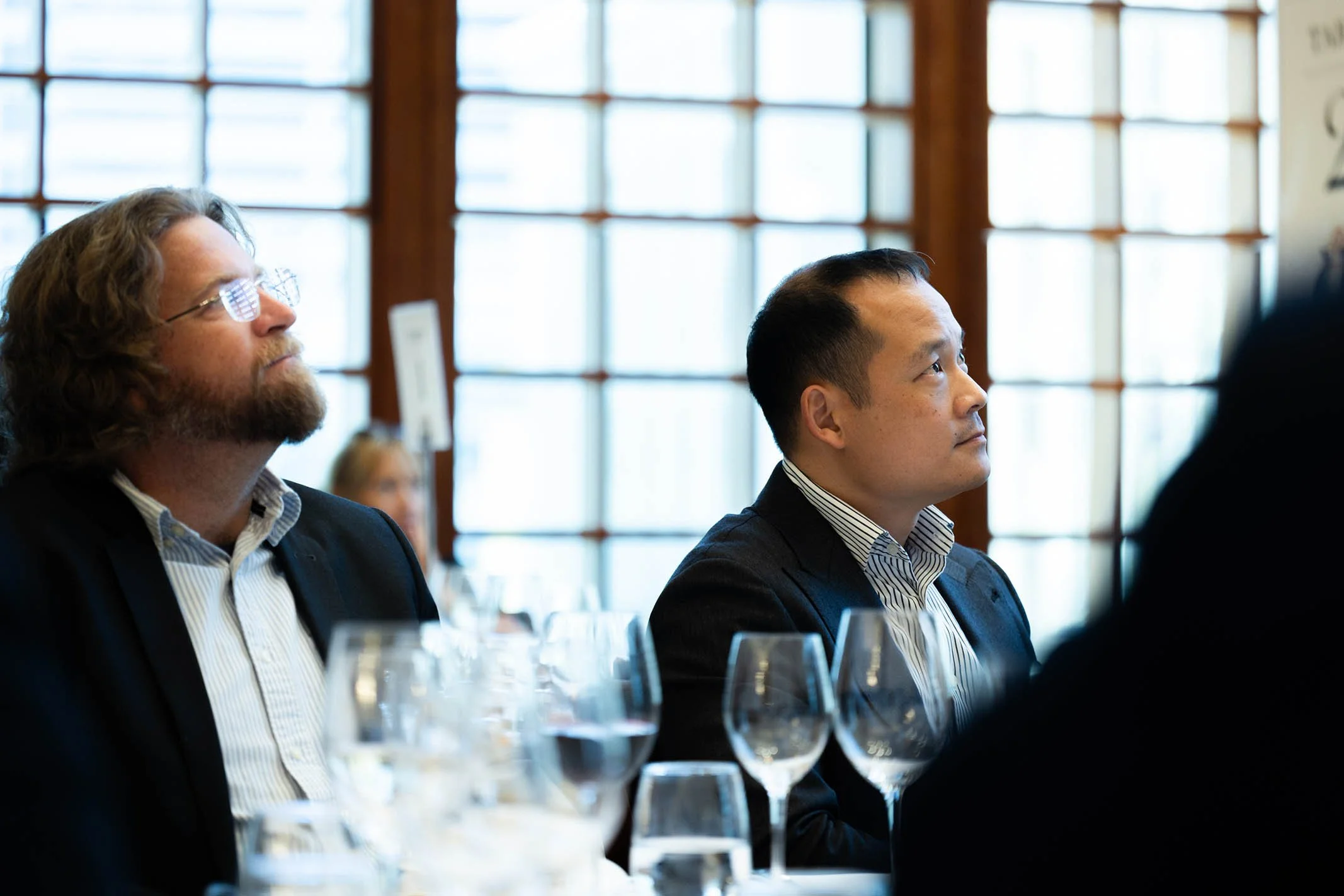 Two men in business attire sitting at a table during a meeting or conference, paying attention to a speaker or presentation, with glasses of water and wine on the table, in a room with large windowpanes.