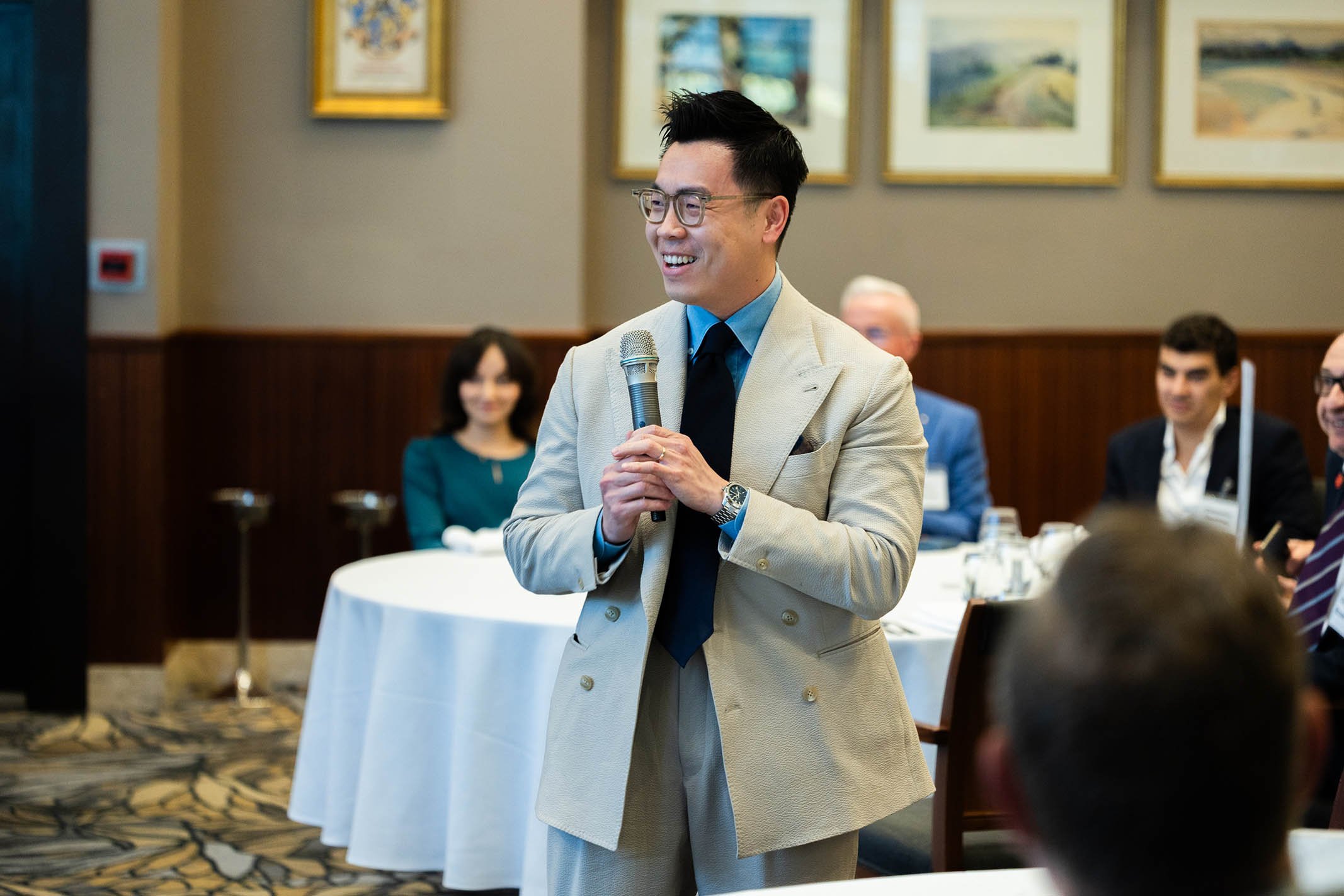 A man in a beige suit holding a microphone, smiling, standing during a formal gathering, with other people seated at round tables in the background inside a decorated conference room.