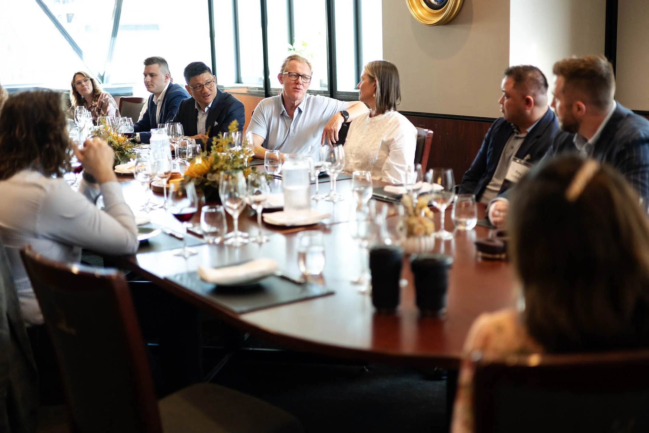 A group of people sitting around a large dining table engaged in conversation at a formal meeting or dinner, with glasses, plates, and floral centerpieces on the table.