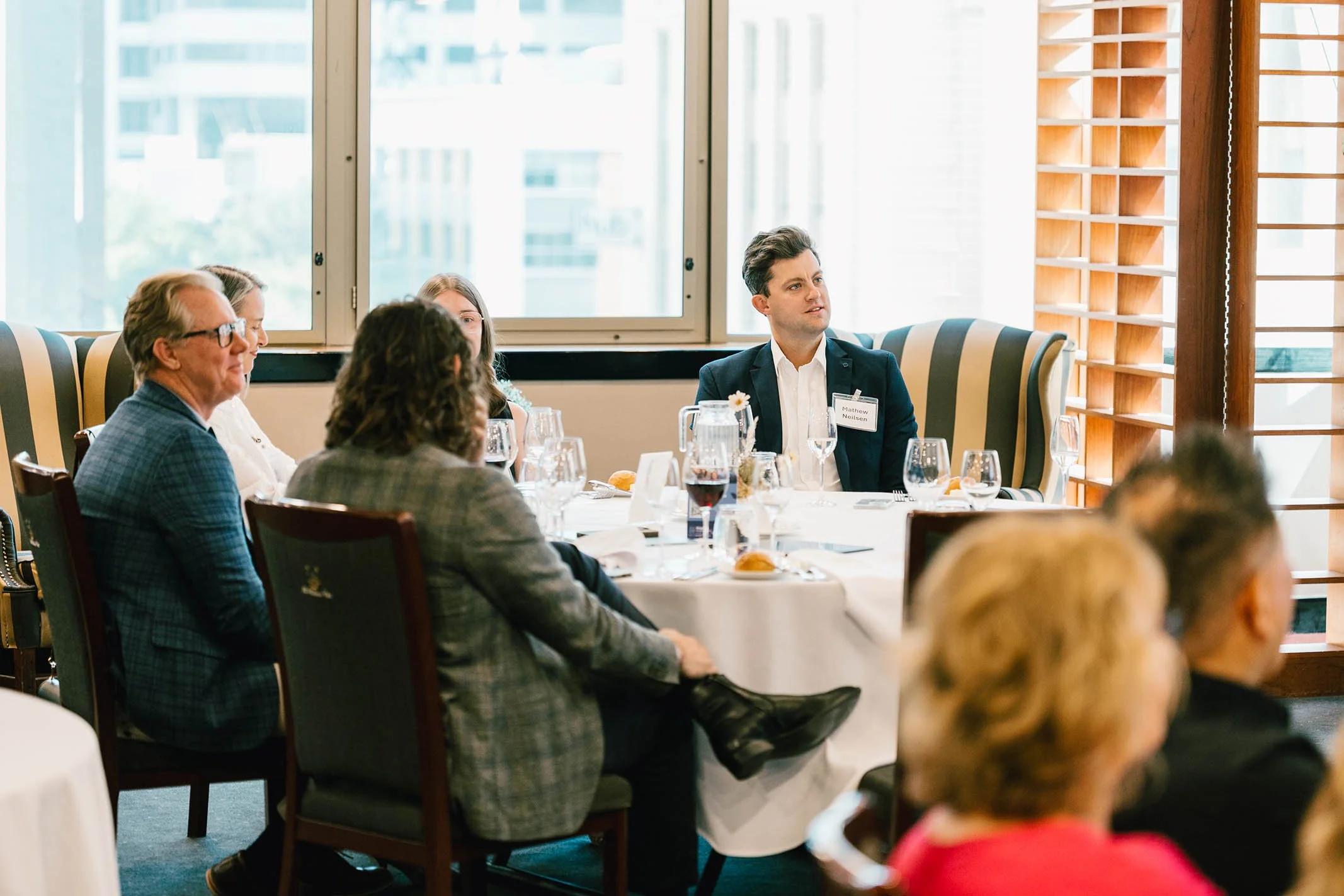 People seated at a round table during a formal event in an indoor room with large windows, some with striped upholstery, and wooden window shutters, engaging in conversation.