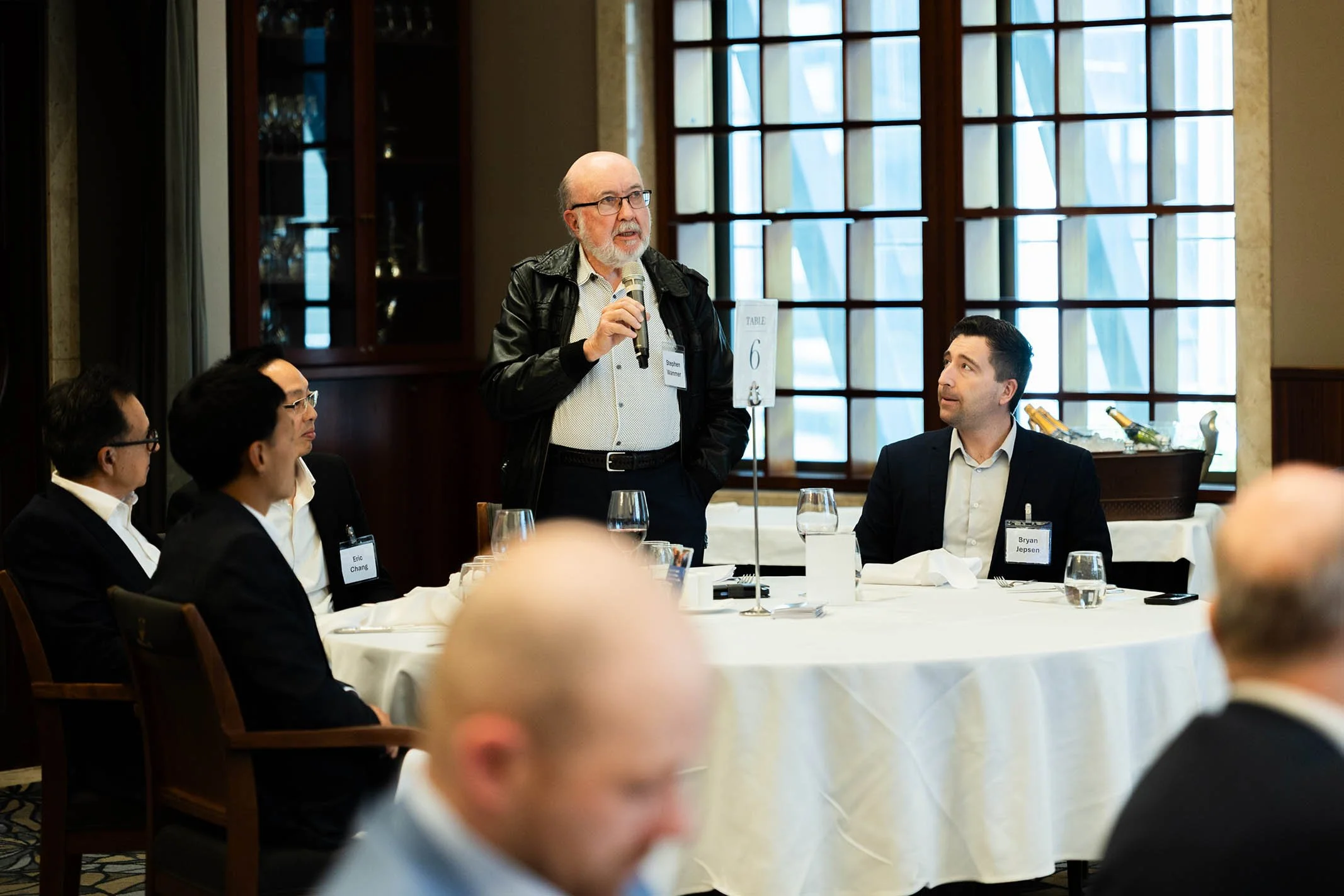 A man with glasses, a white beard, and a black leather jacket is standing and speaking into a microphone at a round table during a conference or meeting. Four other people, three men and one woman, are seated at the table listening. The table has a s