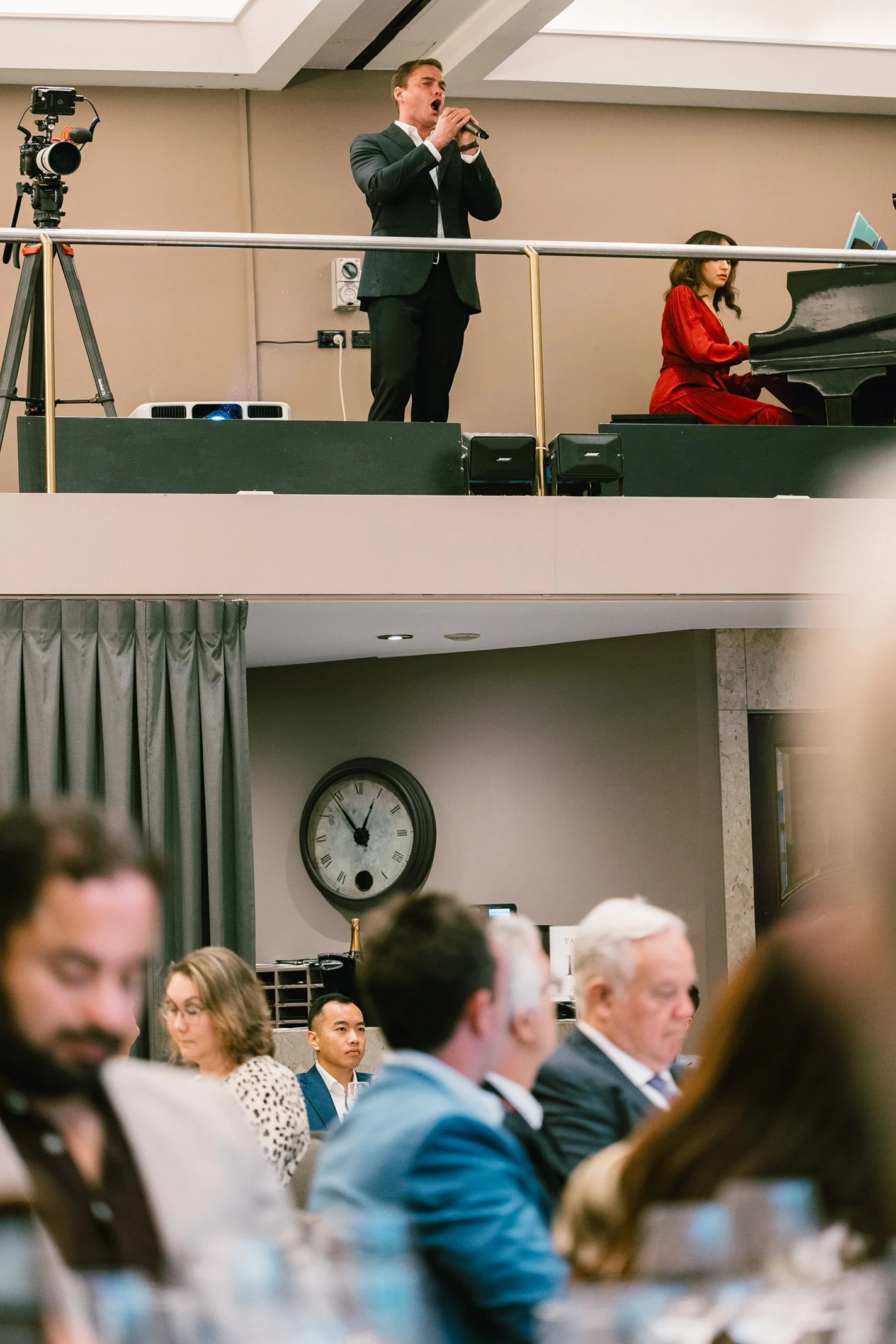 A man in a suit singing into a microphone on an elevated stage, with a woman in a red dress playing the piano beside him, in a room filled with an audience.