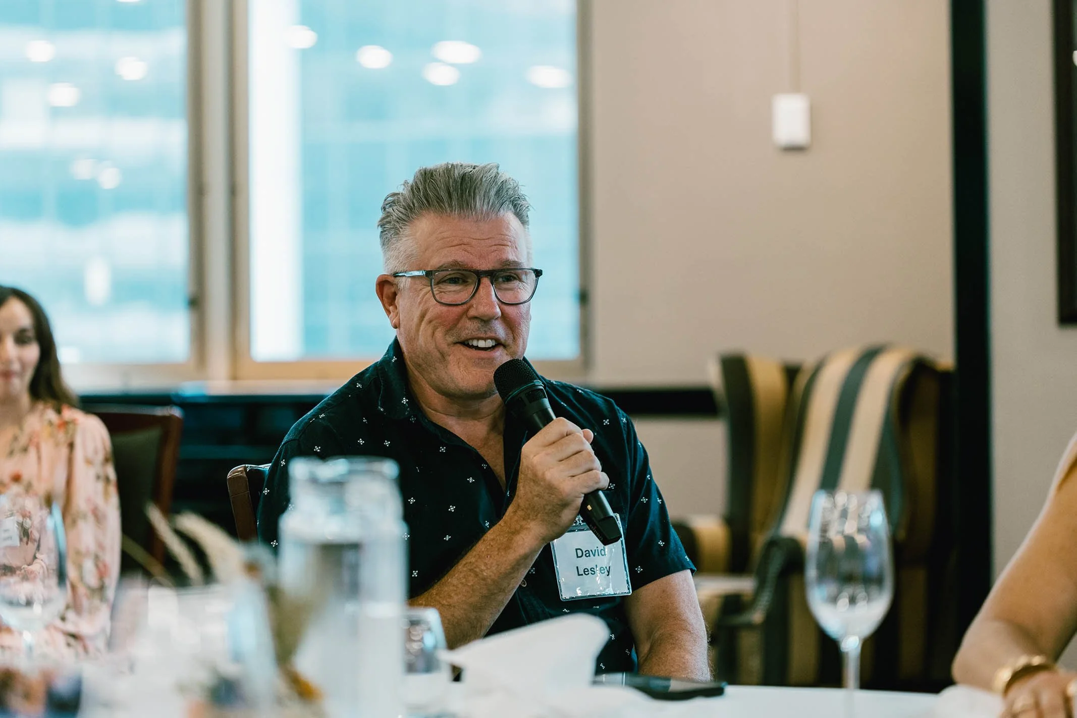 A man with gray hair, glasses, and a black shirt holding a microphone and speaking at a meeting or conference.