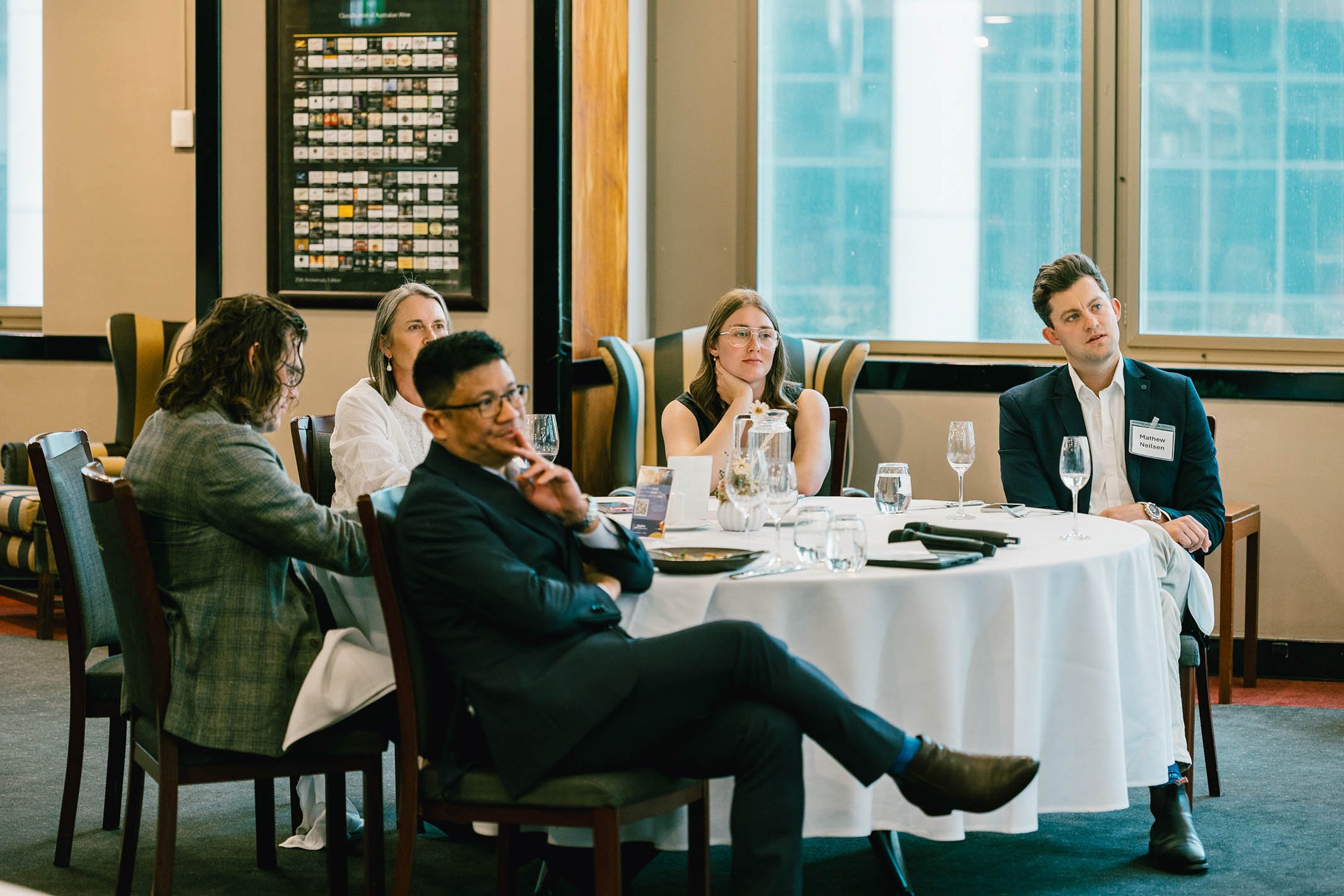 Group of five people sitting at a round table during a formal event or meeting, with large windows in the background and papers and glasses on the table.