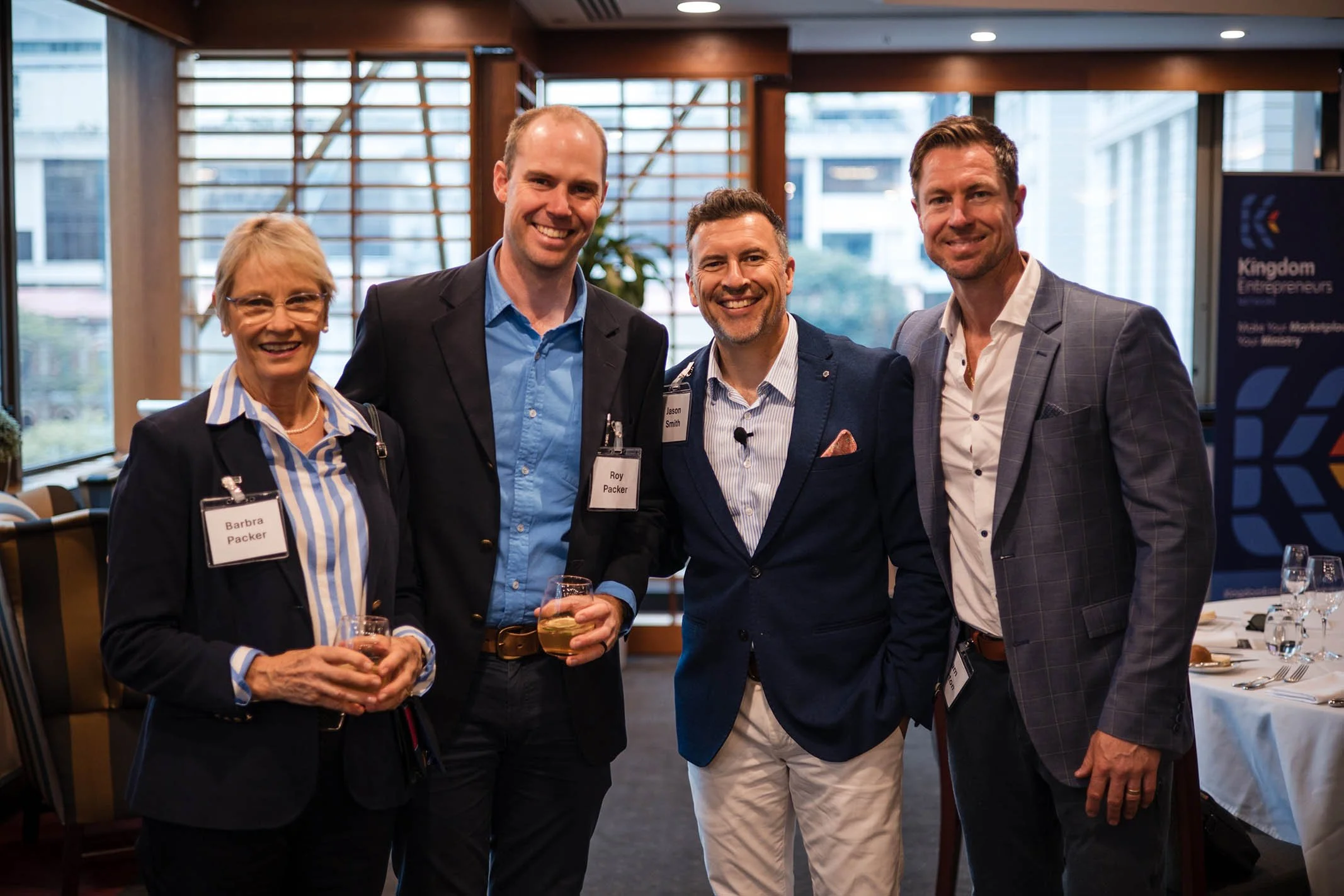 Four professionally dressed individuals standing together at a networking event, smiling at the camera, with name badges visible. The setting appears to be a corporate conference or meeting room with tables and presentation materials in the backgroun