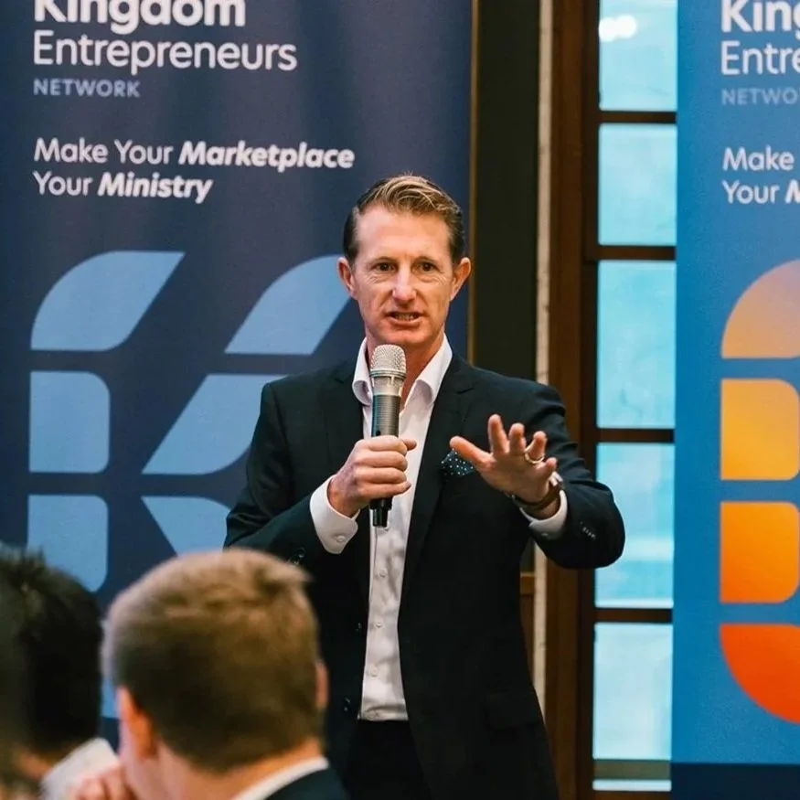 A man in a black suit and white shirt speaking into a microphone at a conference with banners in the background reading "Kingdom Entrepreneurs Network" and "Make Your Marketplace Your Ministry."