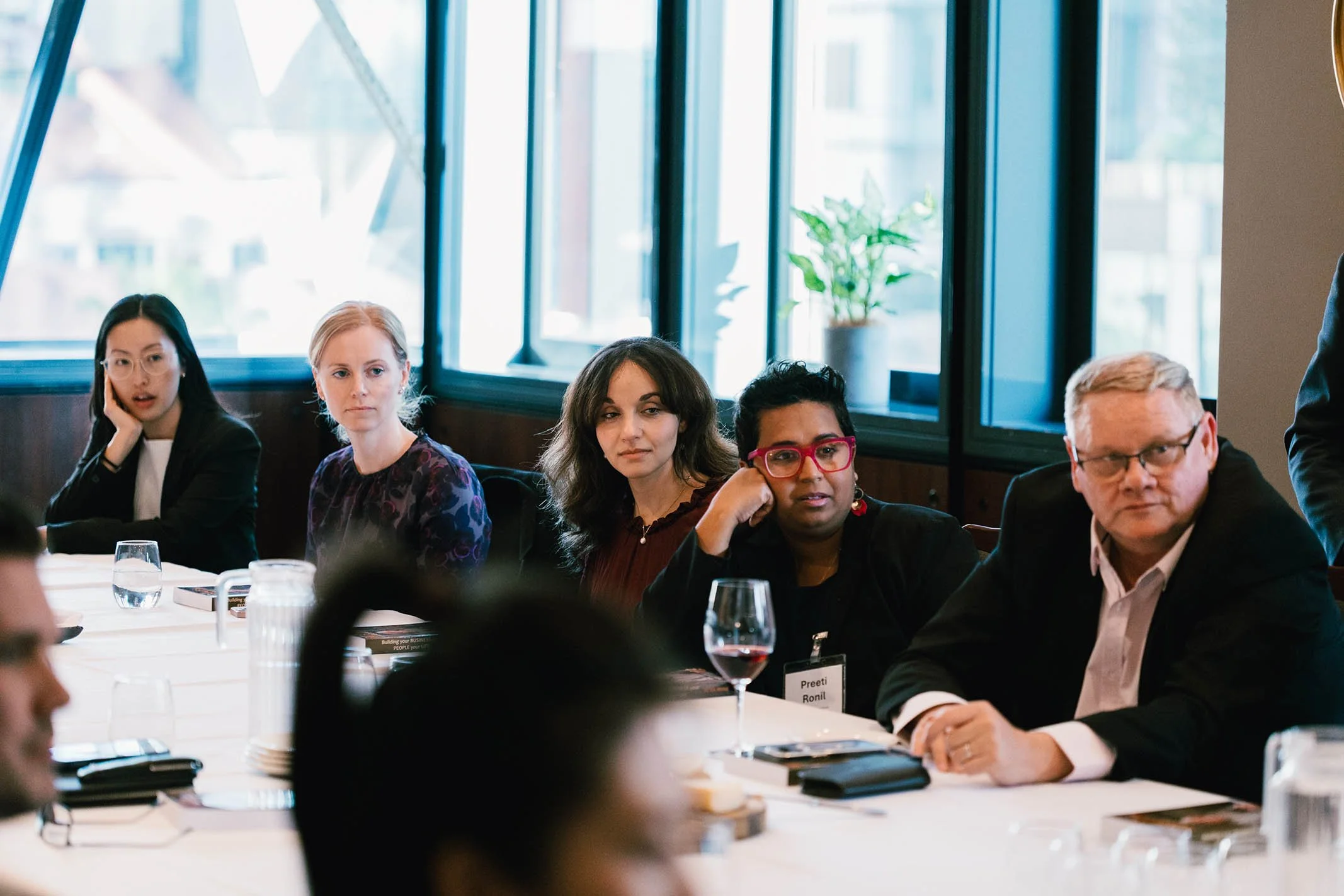 Group of professionals seated at a conference table during a meeting in a bright room with large windows and a potted plant in the background.