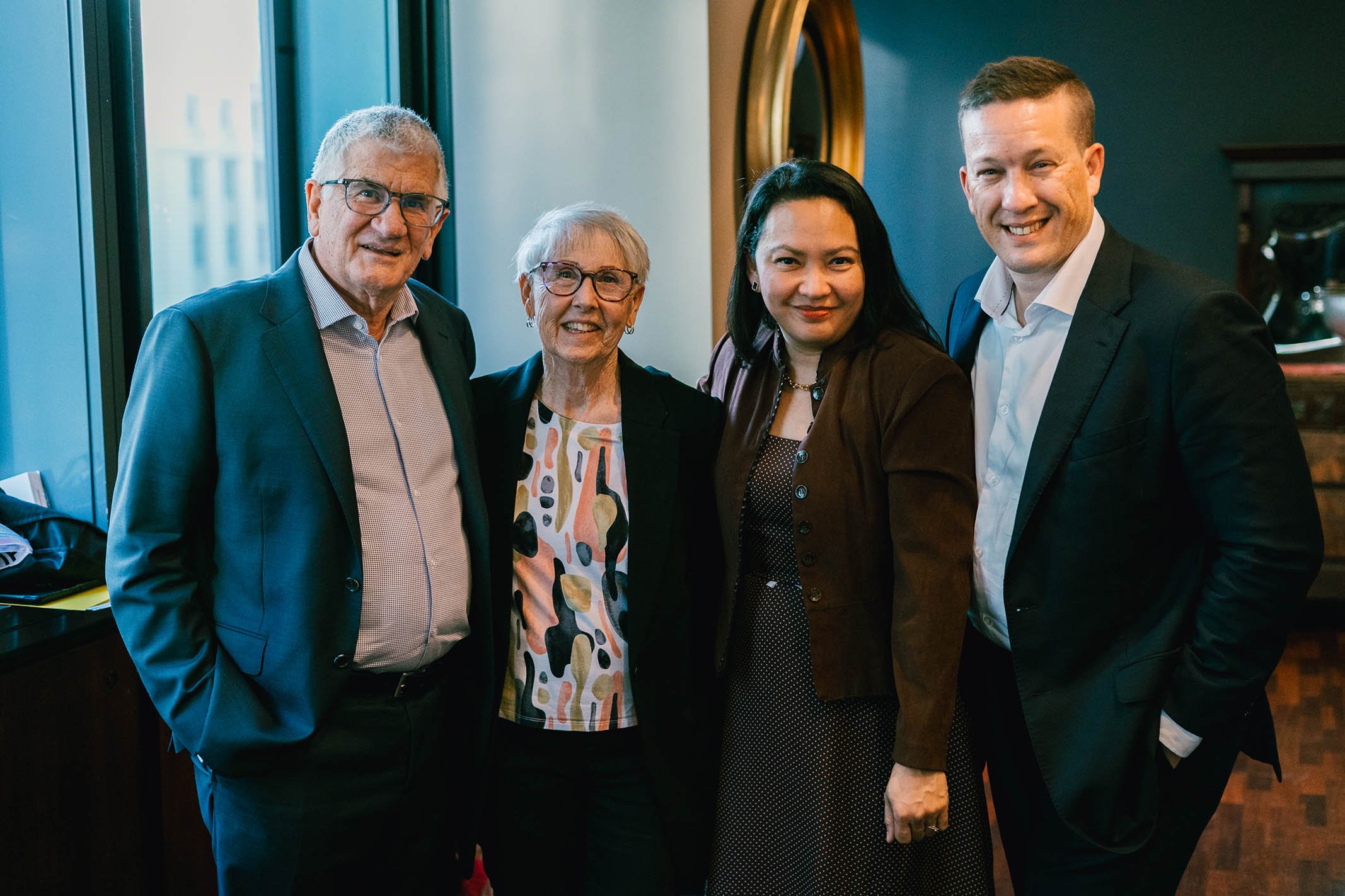 Group of five people, two men and three women, smiling and standing together in a room with dark walls and large windows.