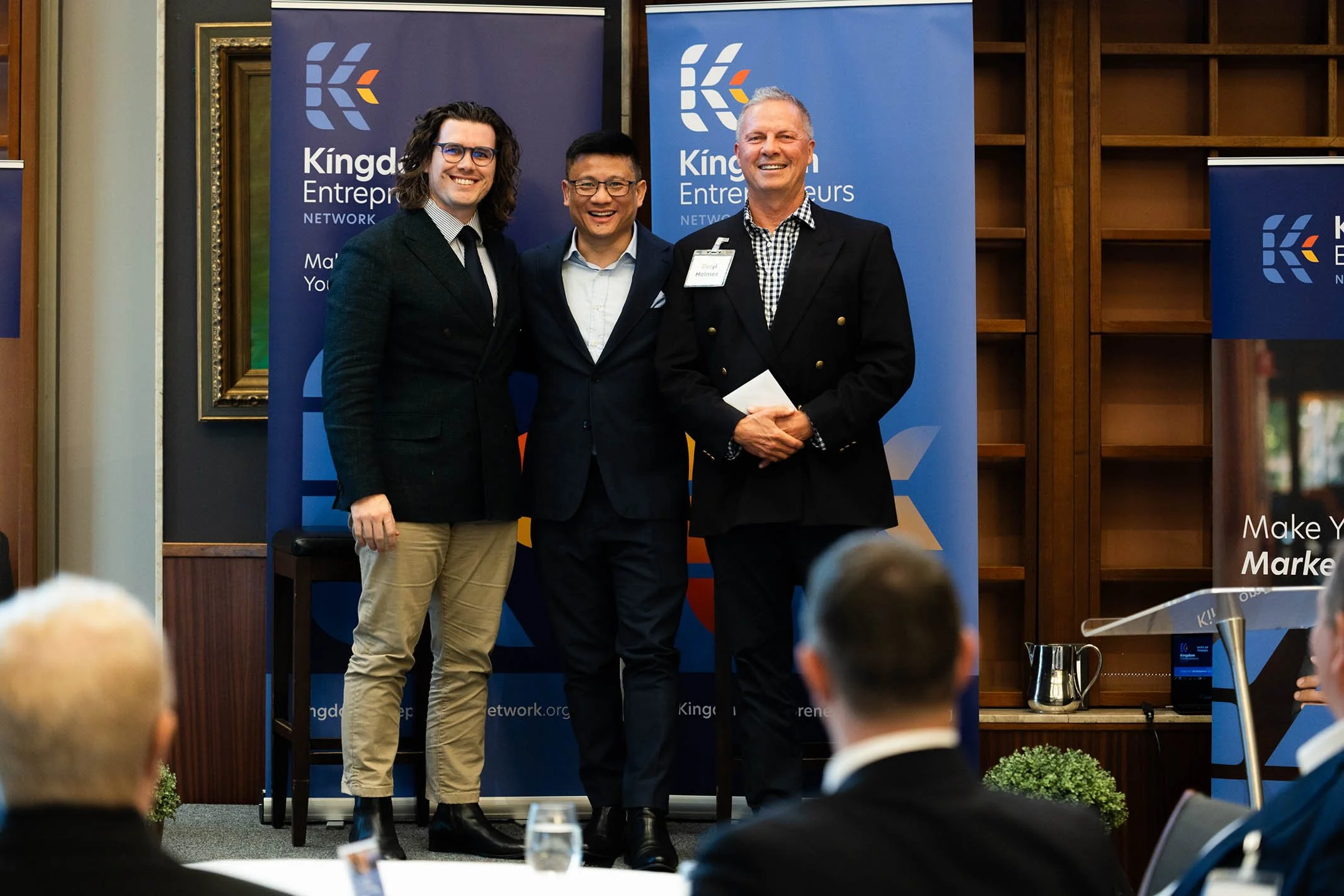 Three men standing together in front of a blue background with 'Kingdom Entrepreneurs' branding at a networking event. All are smiling and dressed in business attire. One man holds a notepad. Audience members are visible seated in the foreground.