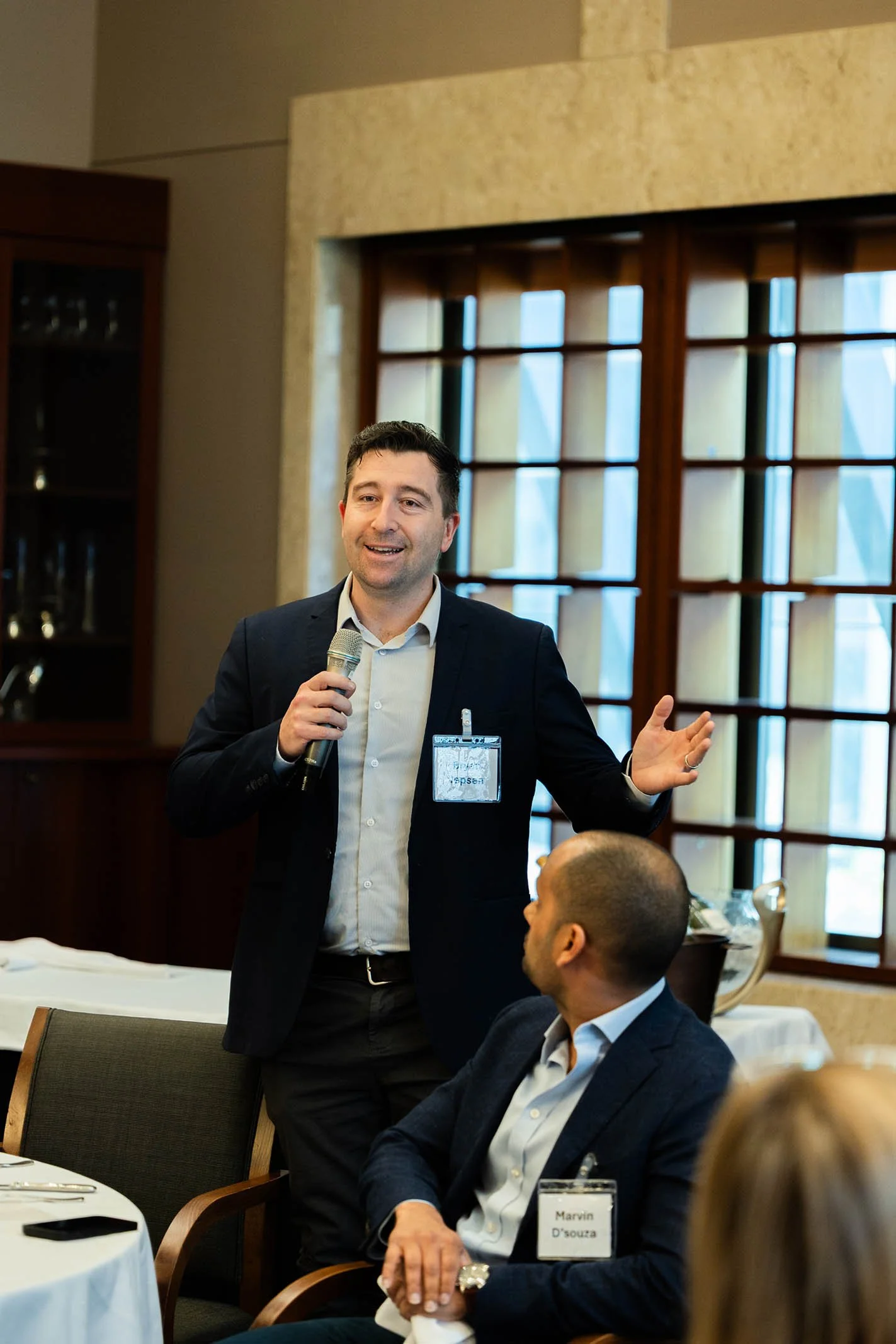 A man in a dark suit and light shirt is speaking into a microphone at a conference or meeting. He is standing next to a seated man with a name badge that reads Marvin D'souza. The setting has large windows with wooden framing and a well-lit interior.
