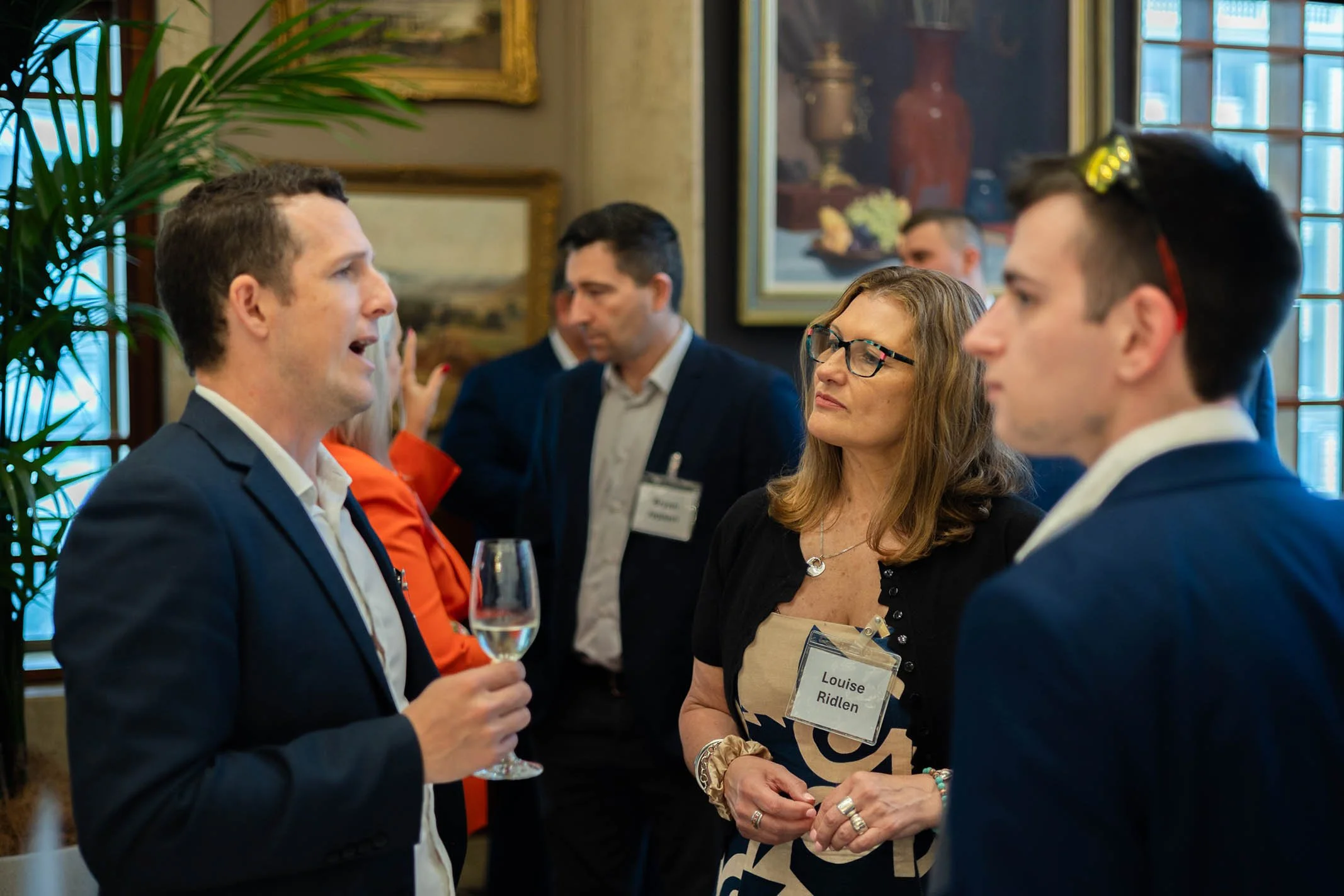 A man in a navy suit is speaking to a woman with glasses, who has a name tag reading 'Louise Ridlen,' during a professional social event with other attendees in the background, some engaging in conversations.
