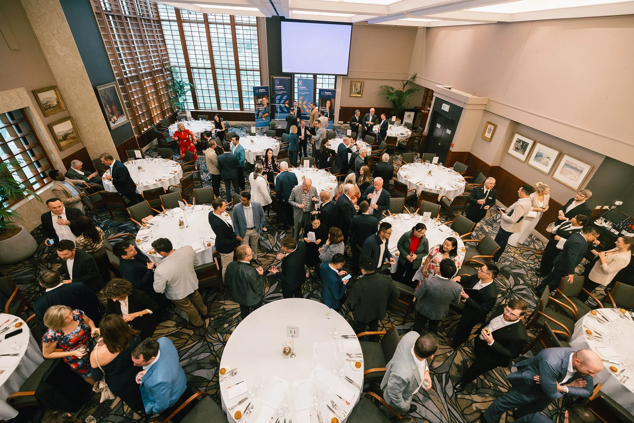 A large indoor event with round tables set for a formal dinner, filled with people socializing and standing in groups, with a stage, large windows, and wall art in the background.
