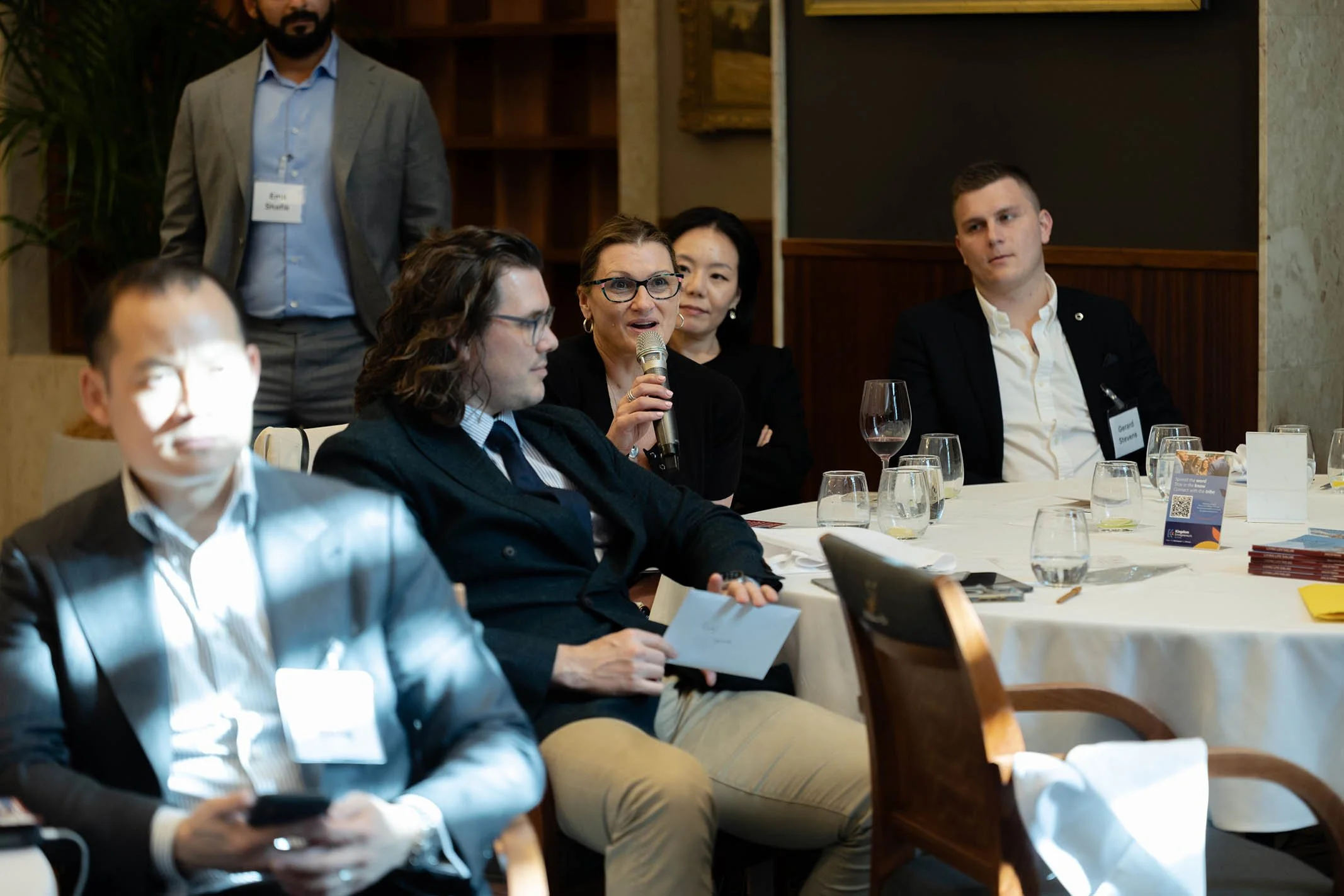 A woman holding a microphone and speaking at a conference, seated at a round table with several other attendees, some with sheets of paper and drinks.