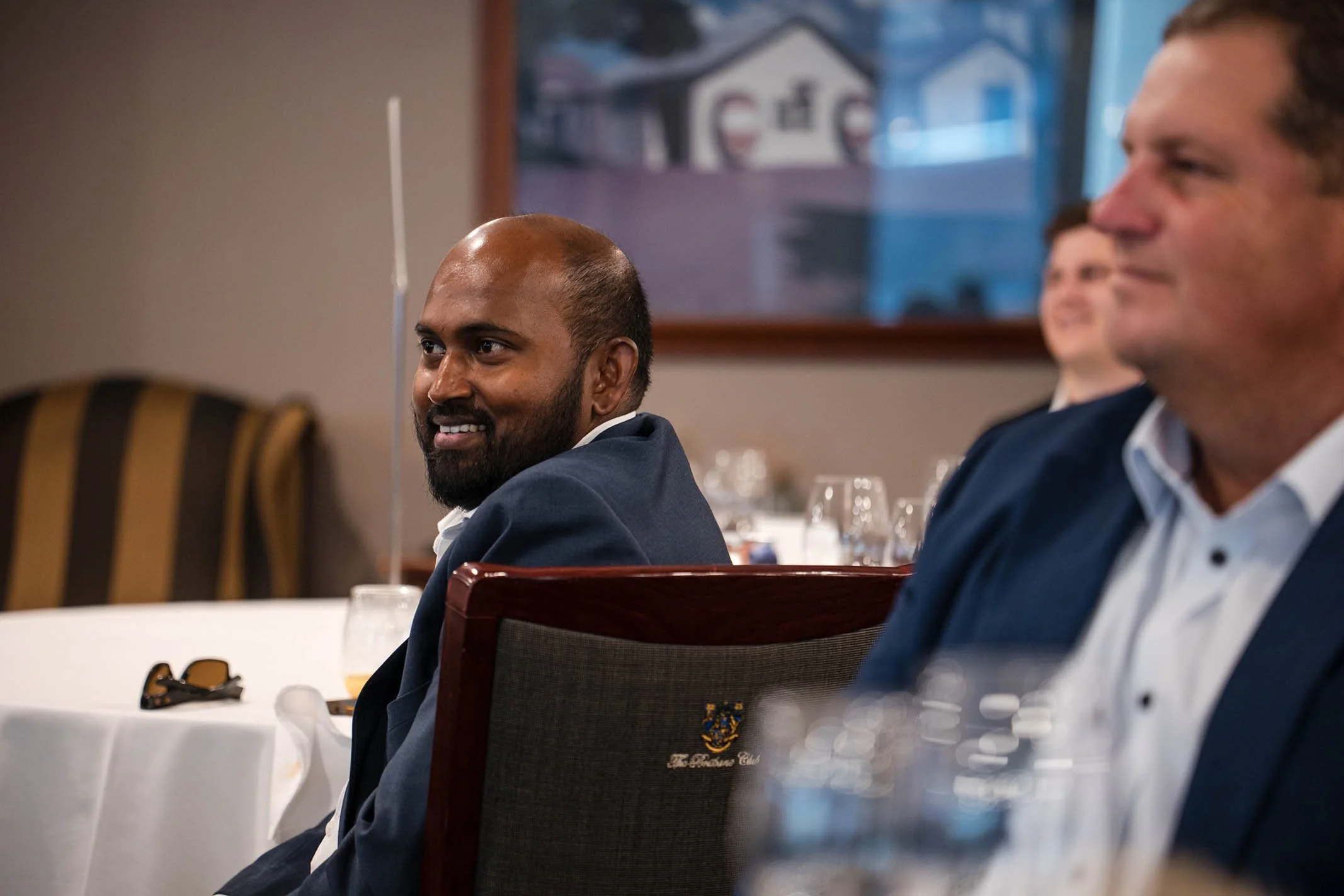 Attendees seated at a formal event, looking towards the front, with tables set with glasses and wine bottles.