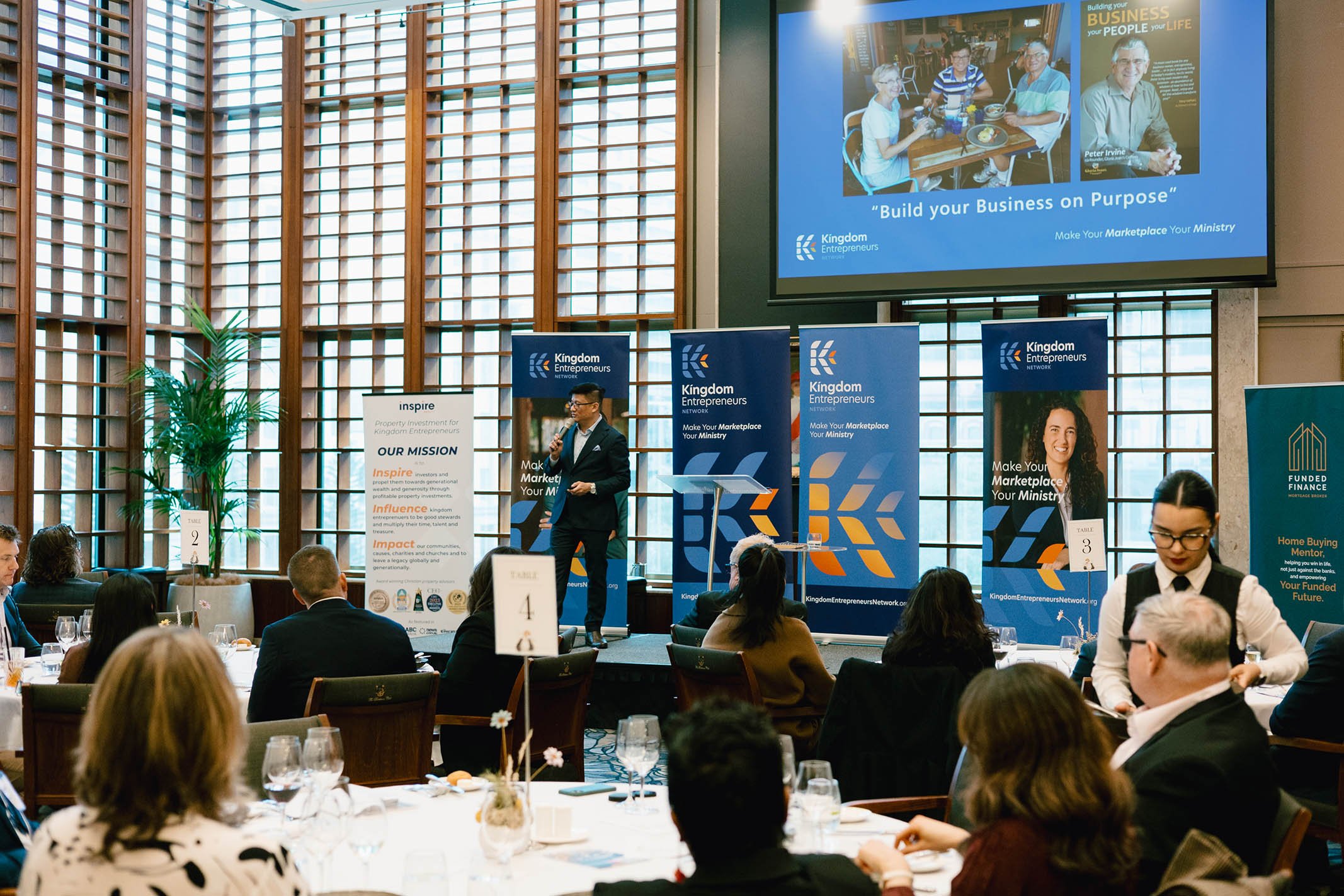 A man speaking on a stage at a conference with banners that say 'Kingdom Entrepreneurs' and 'Build your Business on Purpose.' Audience members seated at tables listen attentively, and a woman in a white shirt and black vest serves at a table.